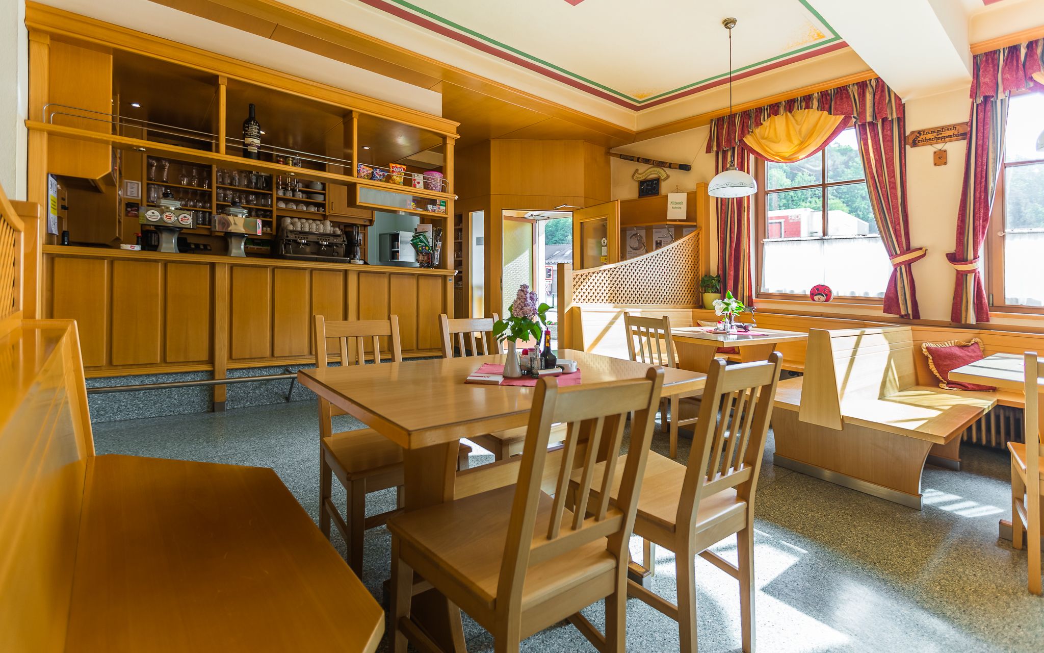 Gemütliches Gastzimmer mit Holzmöbeln, Tischen und Stühlen, dekoriert mit Blumen und Vorhängen, sonnendurchflutet durch große Fenster.