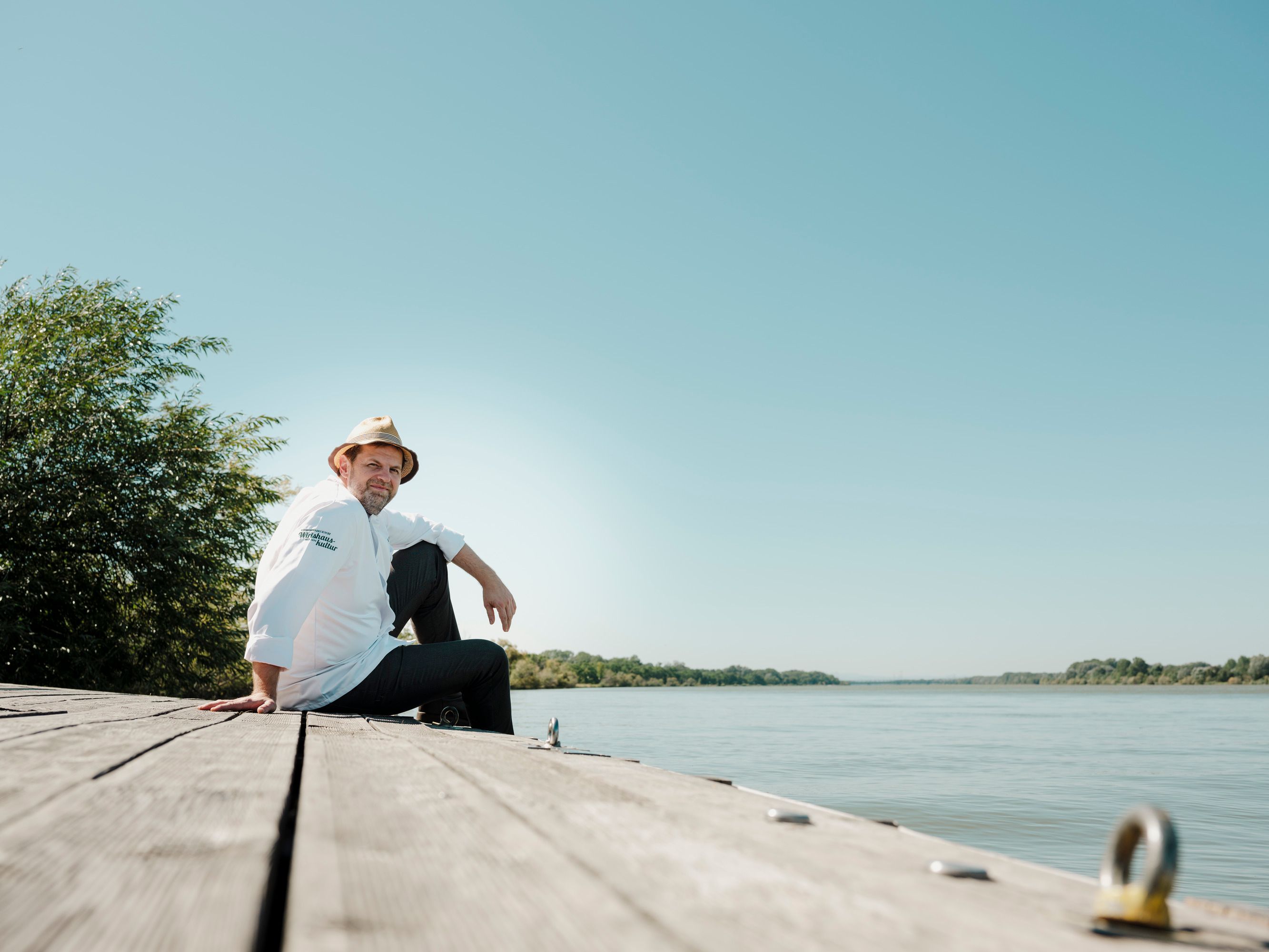 Mann mit Hut und Kochjacke sitzt auf Holzsteg am Flussufer, sonniger Tag.