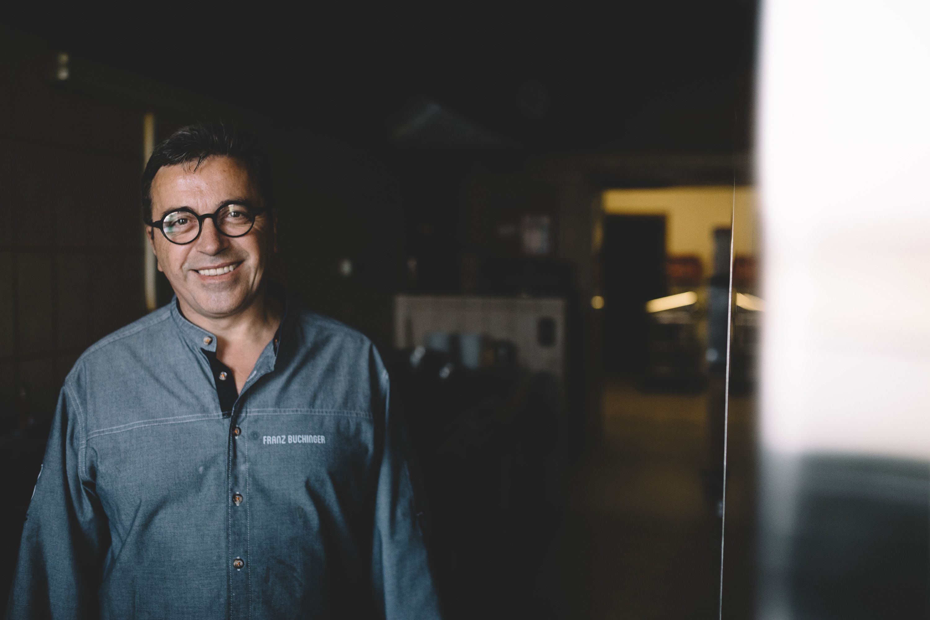 Ein Mann in einer Küche, lächelnd, mit Brille und grauem Hemd.