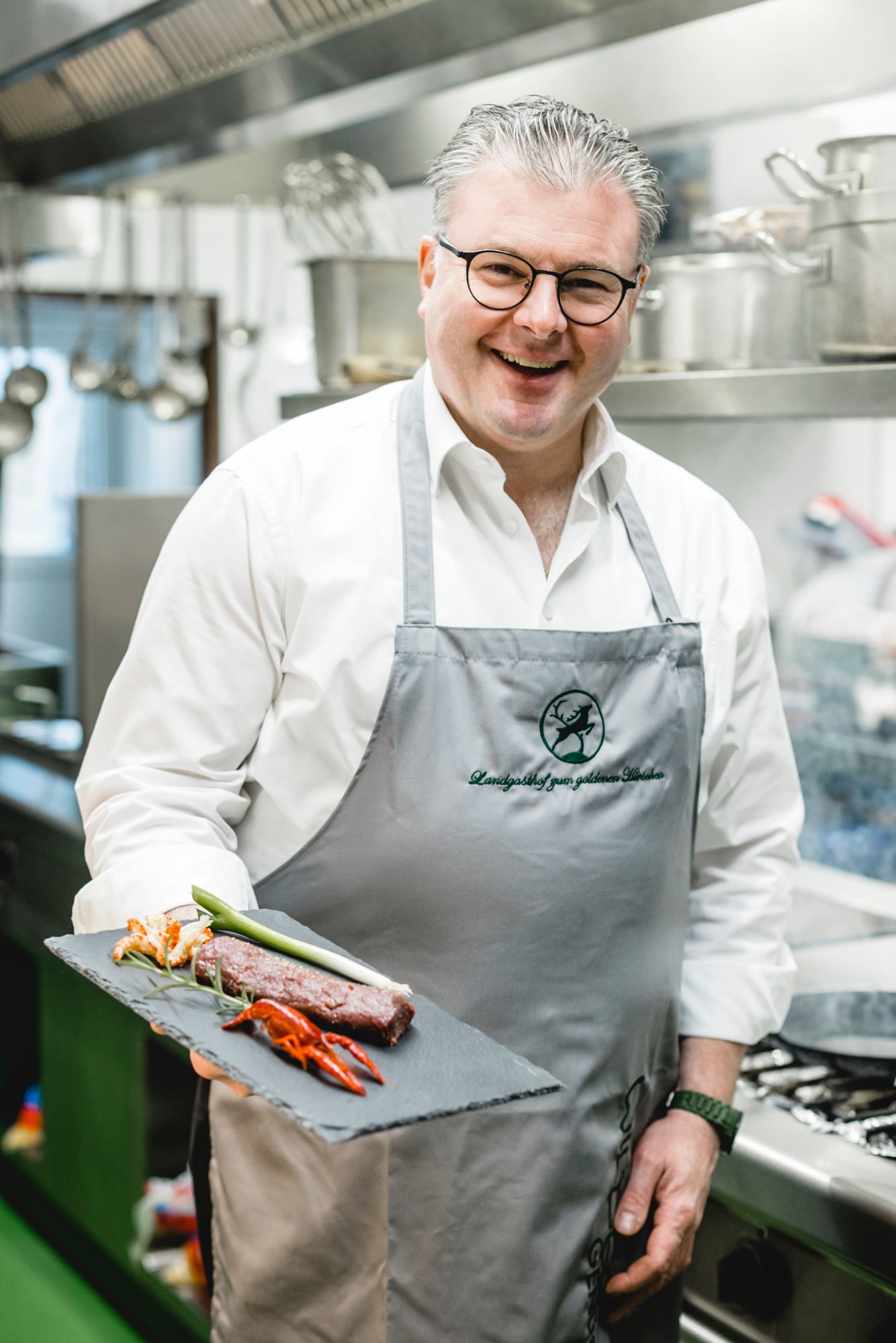 Koch in Küche präsentiert Gericht auf Schieferplatte, lächelt freundlich.