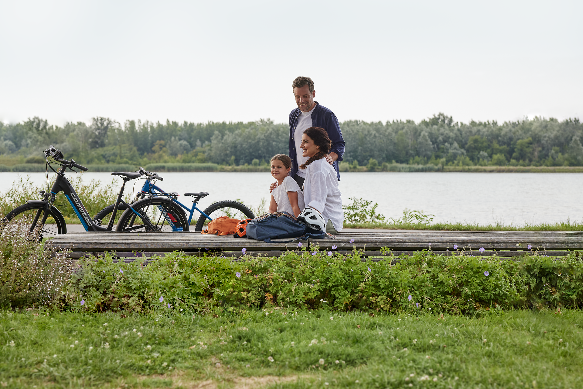 Eine Familie genießt einen entspannten Tag am Ufer der Donau, umgeben von üppigem Grün und blühenden Pflanzen. Die Fahrräder stehen bereit für weitere Erkundungen, während die sanften Wellen des Flusses eine friedliche Atmosphäre schaffen.
