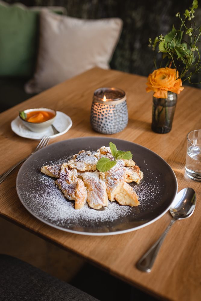 Ein Teller mit Rahmschmarrn, bestreut mit Puderzucker, auf einem Holztisch. Daneben eine Schale mit Marillenröster und eine brennende Kerze.