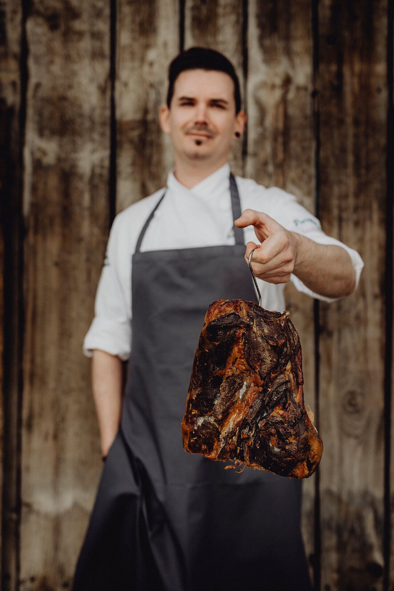 Ein leidenschaftlicher Koch präsentiert stolz ein perfekt zubereitetes Stück Fleisch, das die Tradition der Wirtshauskultur verkörpert. Die rustikale Holzverkleidung im Hintergrund schafft eine einladende Atmosphäre, die zum Verweilen einlädt und die Sinne für die kulinarischen Genüsse der Region öffnet.