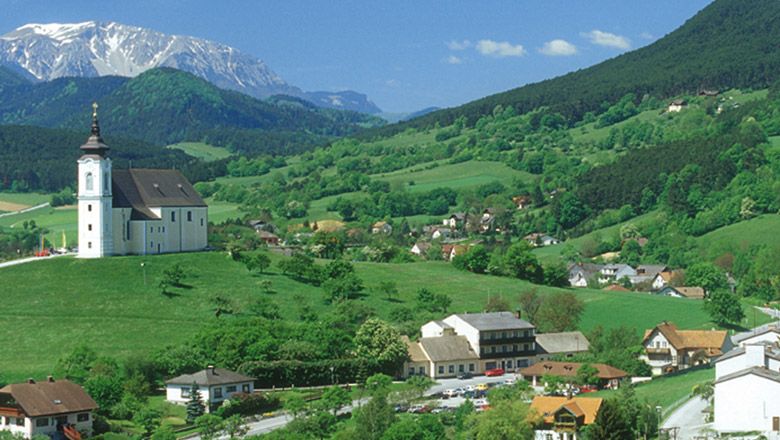 Landschaft mit Kirche auf Hügel, umgeben von grünen Feldern und Bergen im Hintergrund.
