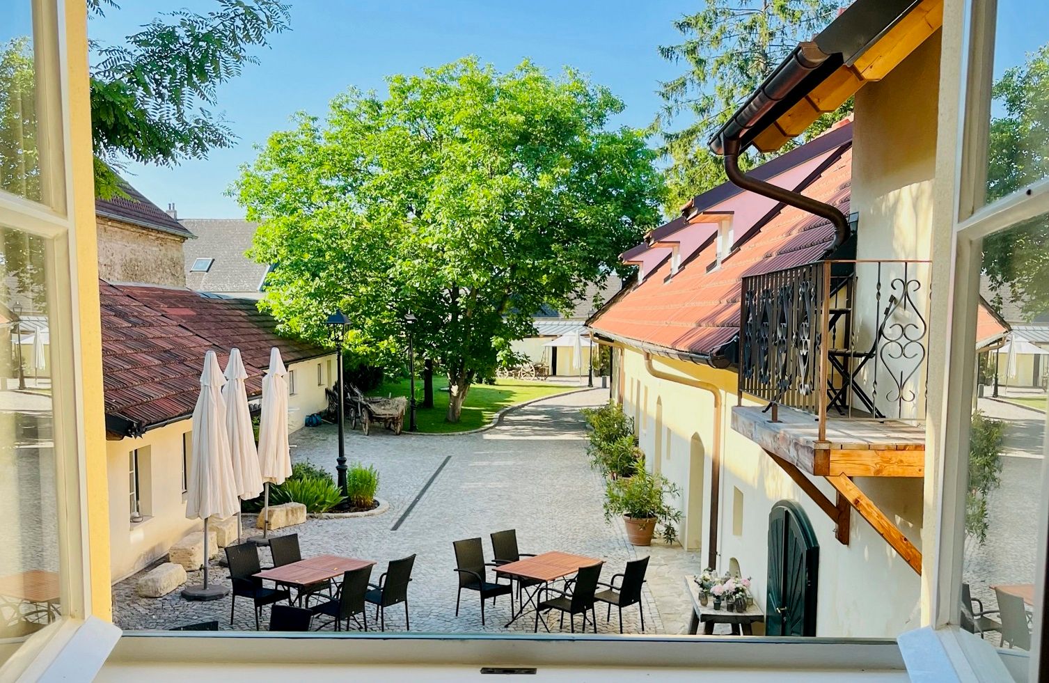 Blick aus einem Fenster auf einen Innenhof mit Tischen, Stühlen und Sonnenschirmen, umgeben von Gebäuden und Bäumen.