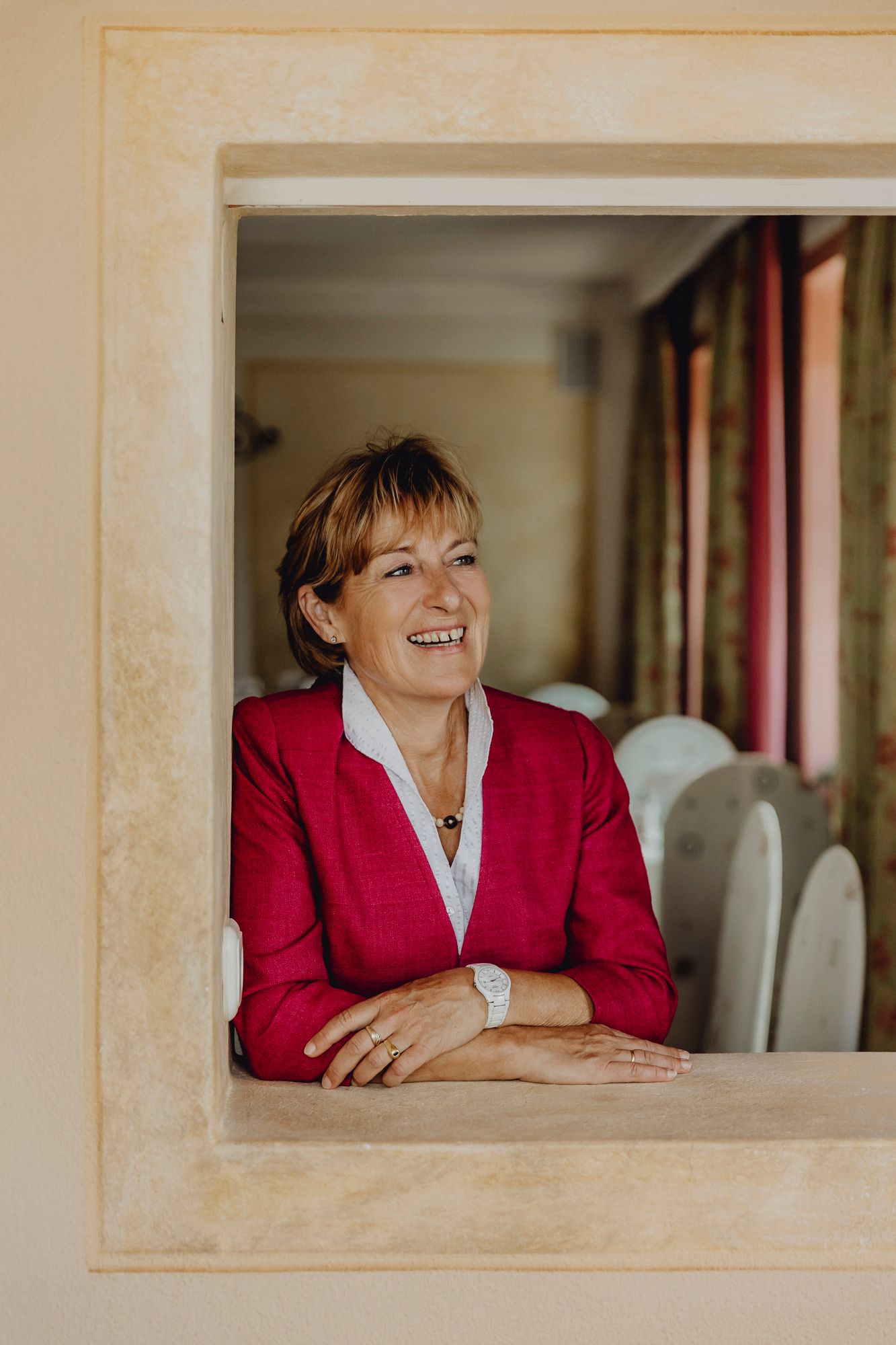 Eine lächelnde Frau in einem roten Blazer lehnt sich durch ein Fenster.