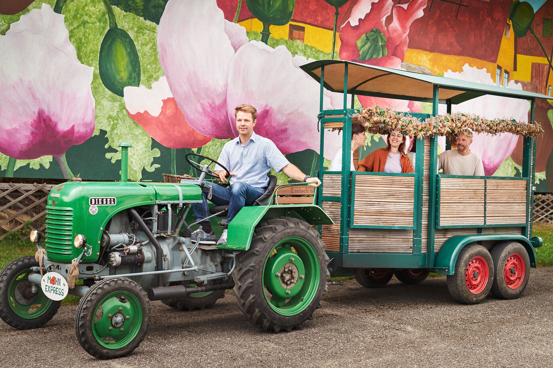 Inmitten der blühenden Mohnfelder strahlt die ländliche Idylle. Ein nostalgischer Traktor zieht eine liebevoll dekorierte Wagenladung, während die sanften Hügel im Hintergrund die Schönheit der Natur unterstreichen. Hier erleben Besucher die Faszination des Mohns und die herzliche Gastfreundschaft der Region.
