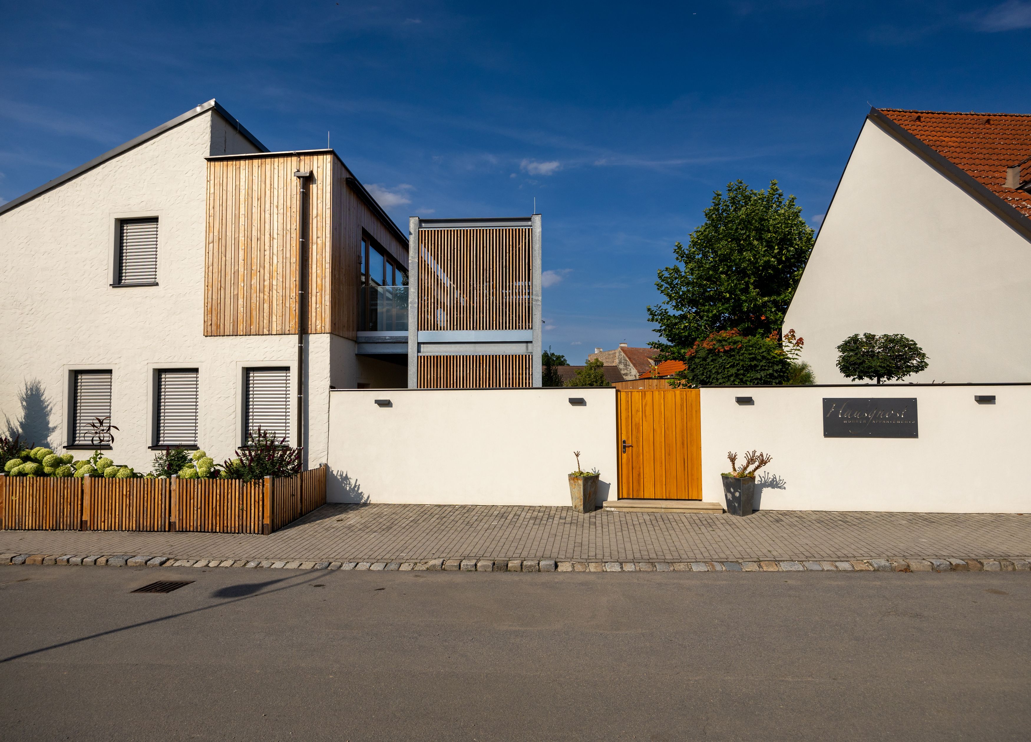 Modern building with a white façade and wooden elements, surrounded by a wall with a wooden gate.