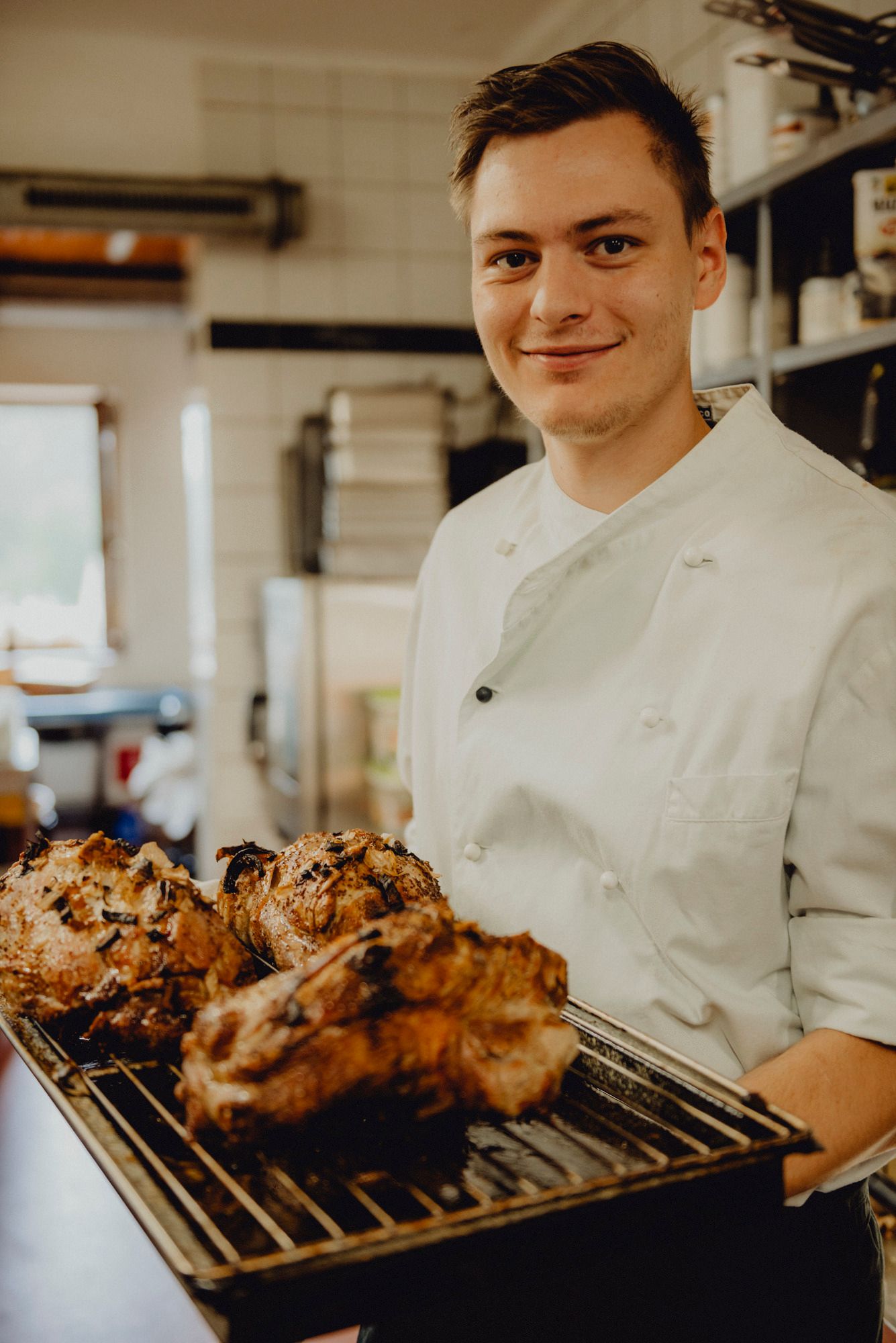 Ein Koch in weißer Uniform hält ein Tablett mit gebratenem Fleisch in einer Küche.