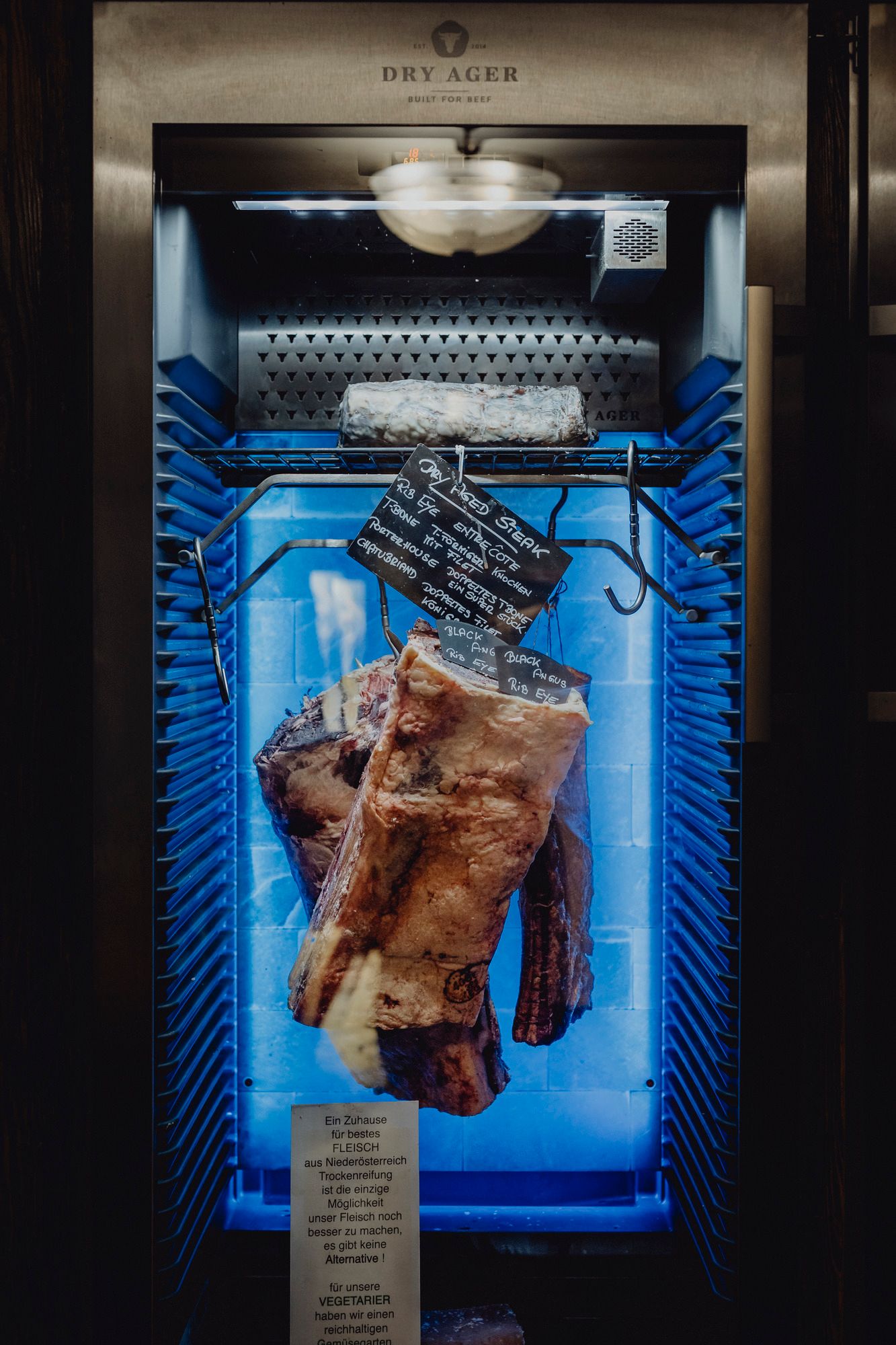 Ein Dry Ager Kühlschrank mit hängendem Fleisch und einer Tafel mit Informationen.