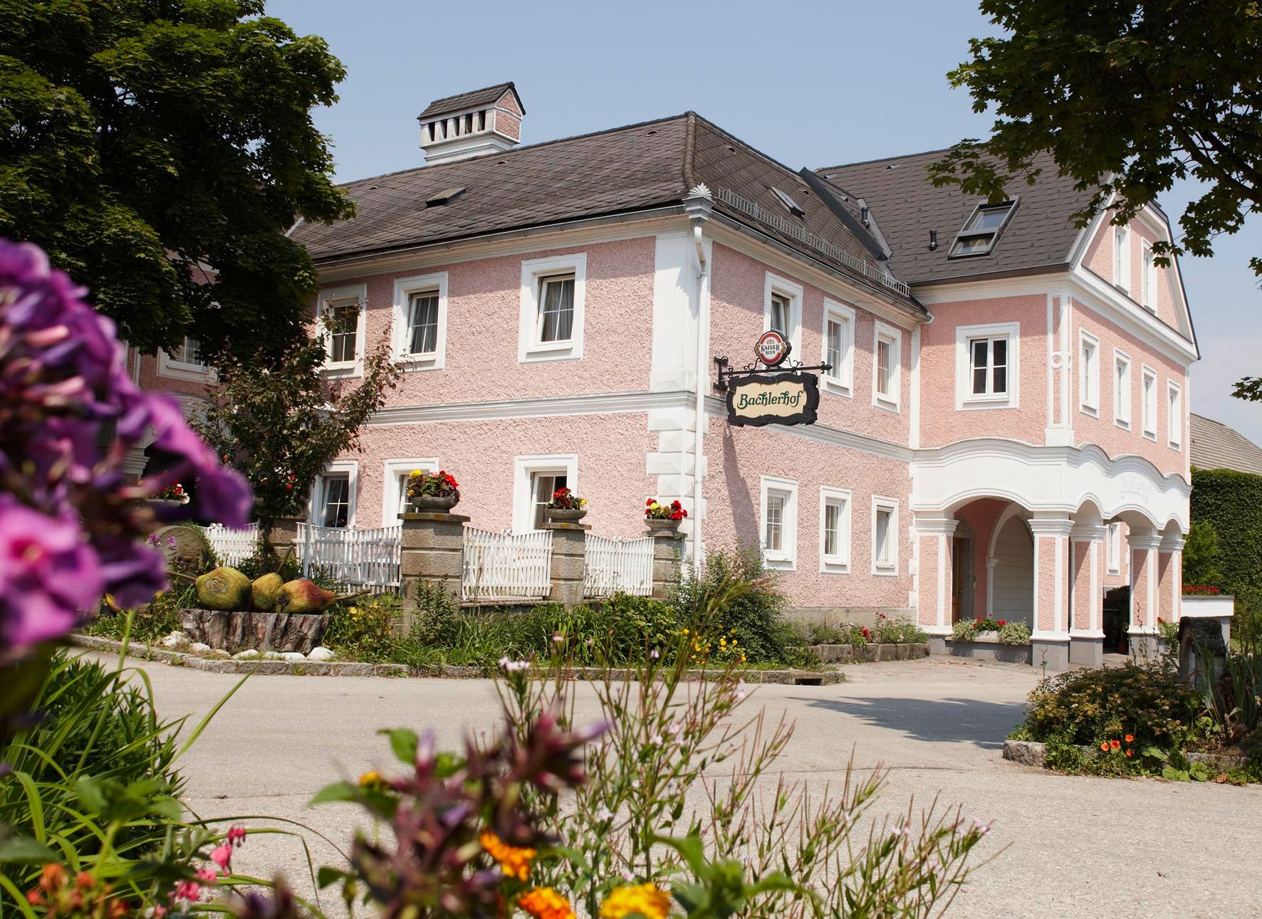 Ein traditionelles, rosa gestrichenes Gebäude mit dem Schild 'Bachlerhof', umgeben von Blumen und Bäumen.
