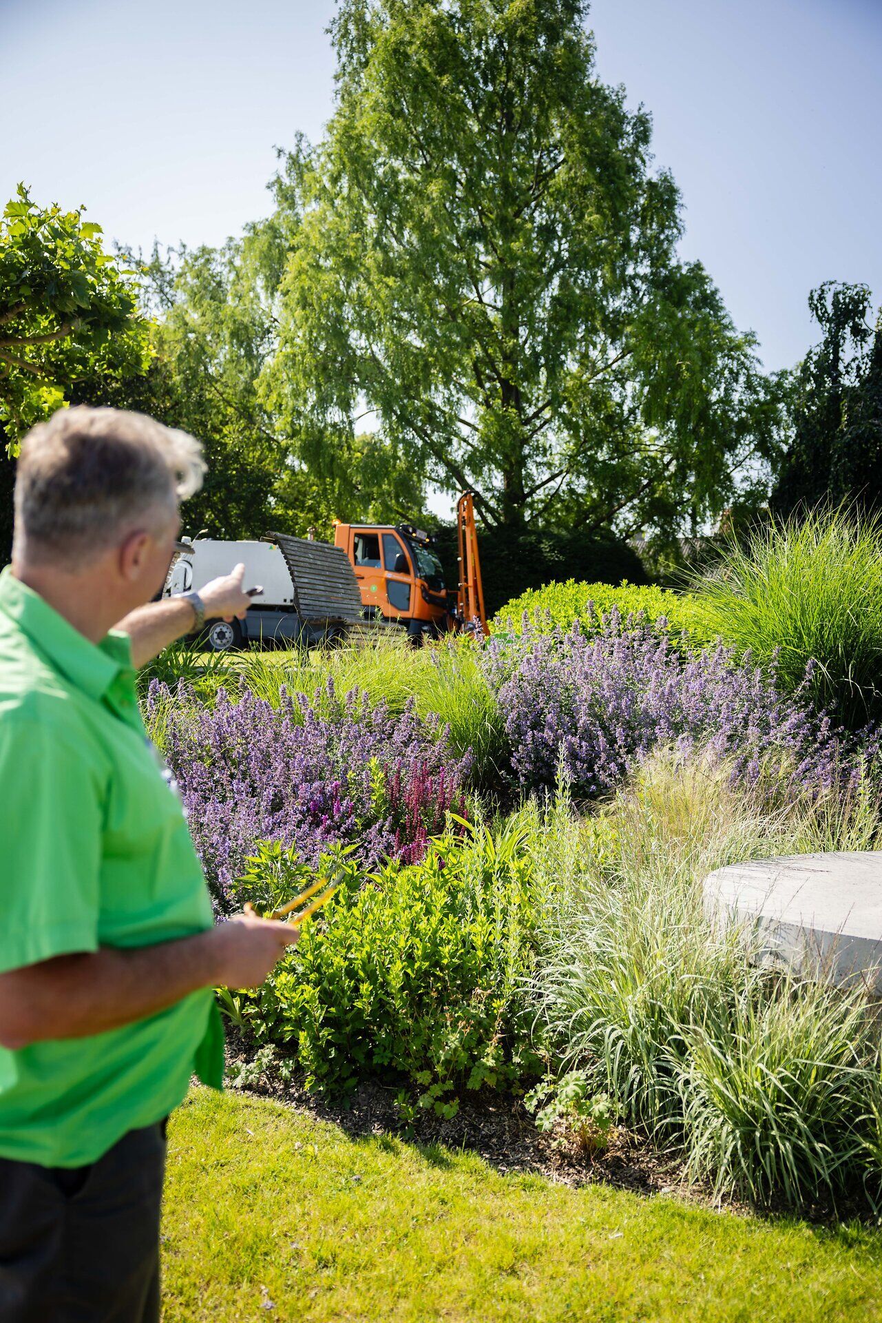 Ein Gärtner in Tulln zeigt auf einen Baum, davor steht ein Einsatzfahrzeig, überall sind blühende Sträucher und Bäume zu sehen, blauer Himmel und Sonnenschein.