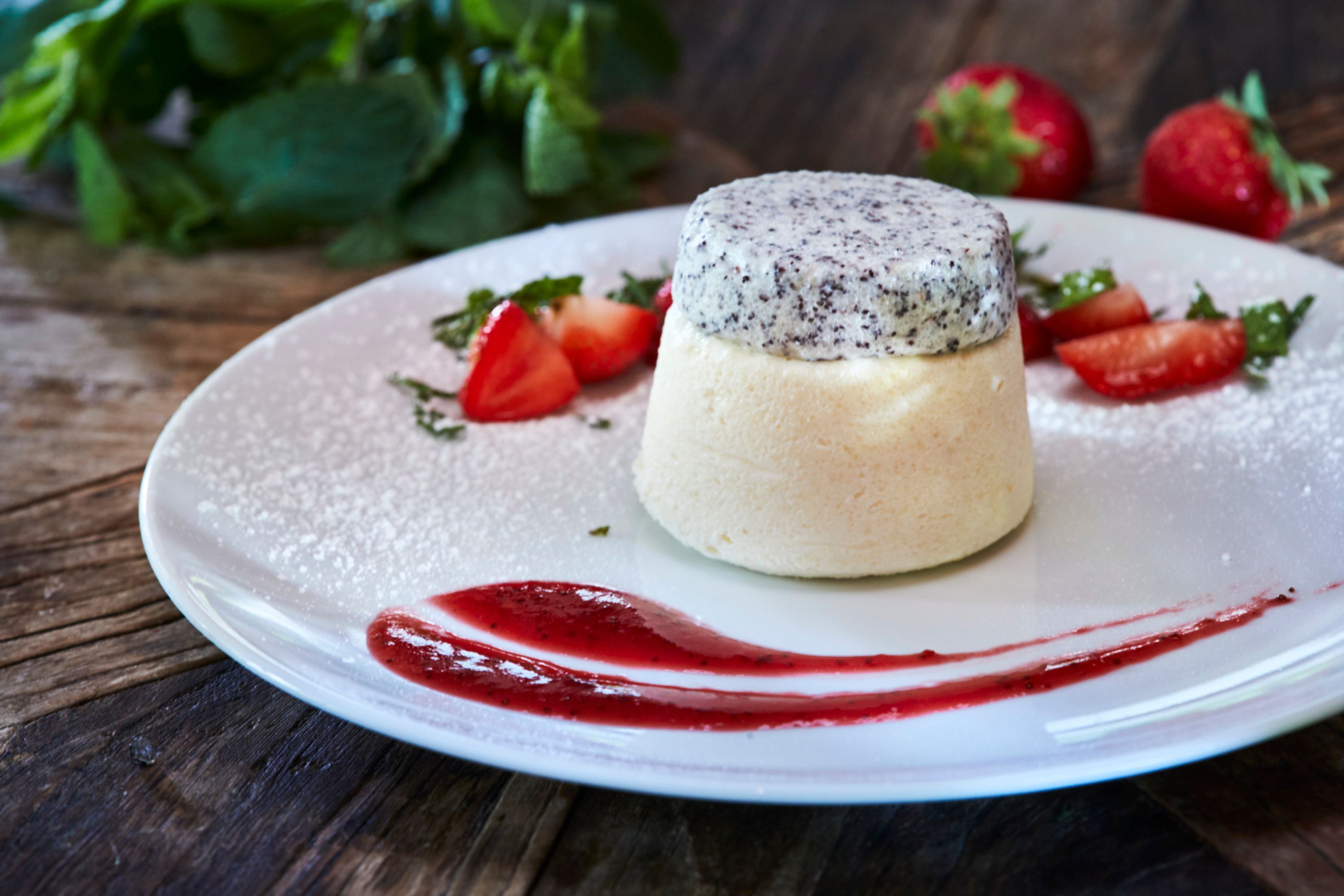 Ein Dessert aus Grießflammerie mit Mohnhaube, garniert mit Erdbeeren und Minze auf einem weißen Teller.