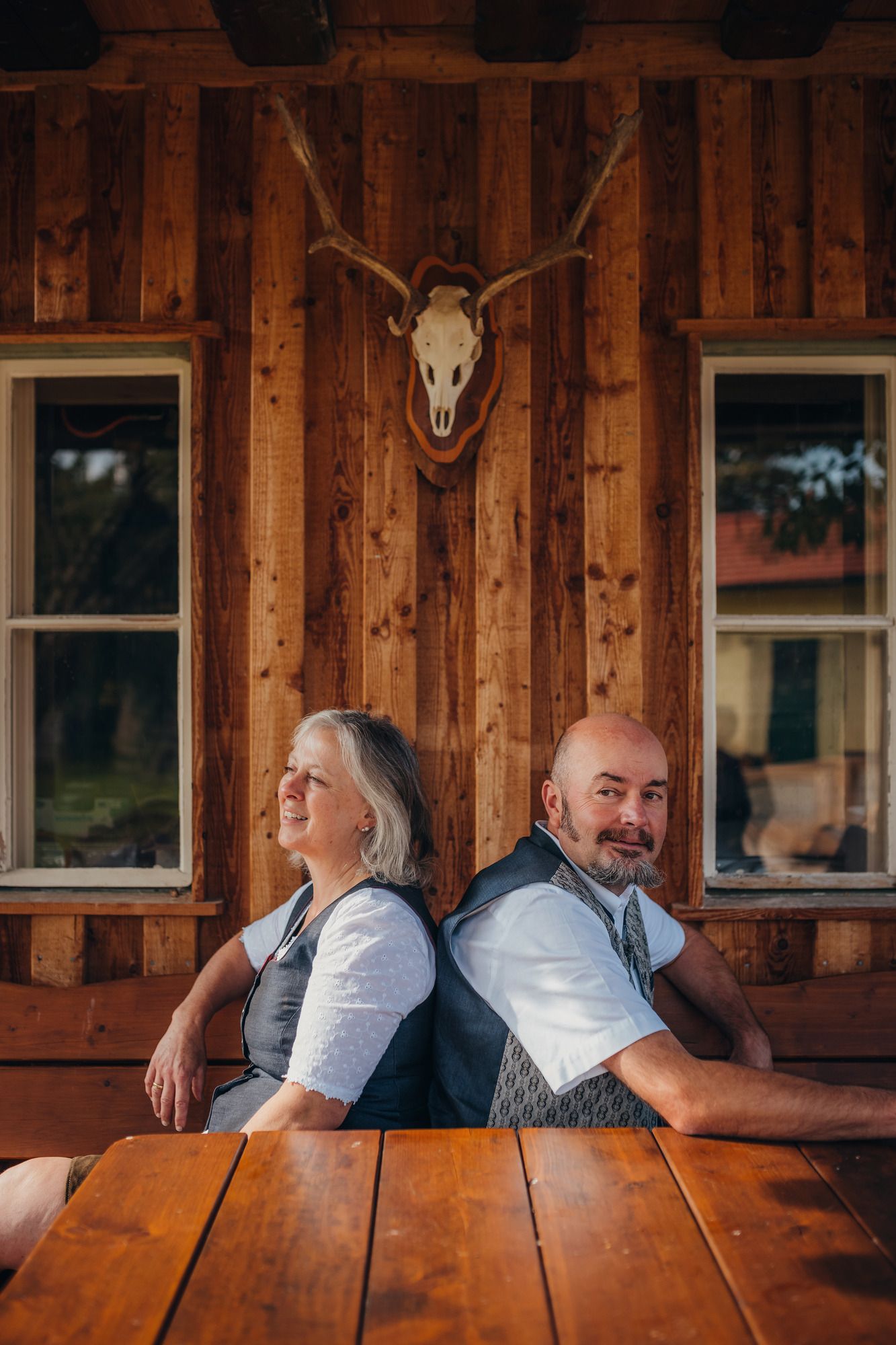 Ein Mann und eine Frau sitzen Rücken an Rücken vor einer Holzwand mit einem Hirschgeweih.