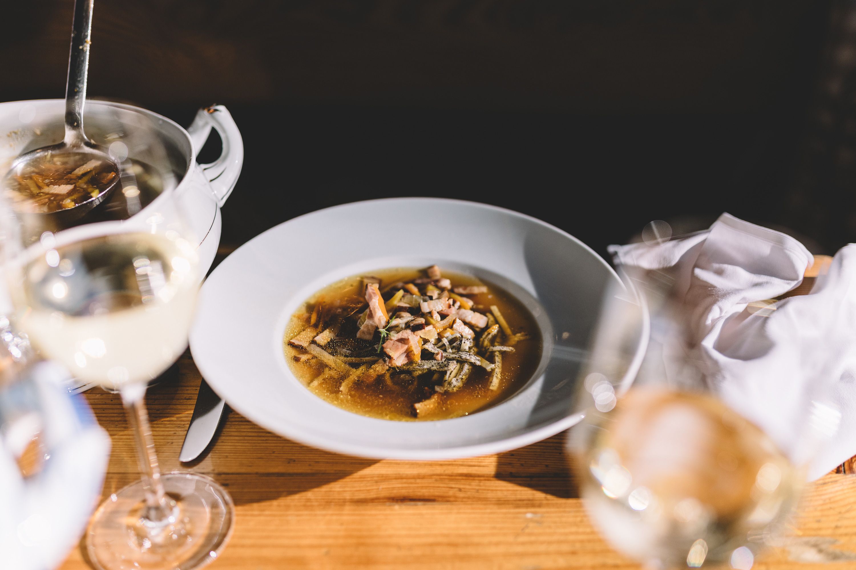 Ein Teller mit Roggenbiersuppe und Mohnfrittaten auf einem Holztisch, daneben ein Glas Weißwein.
