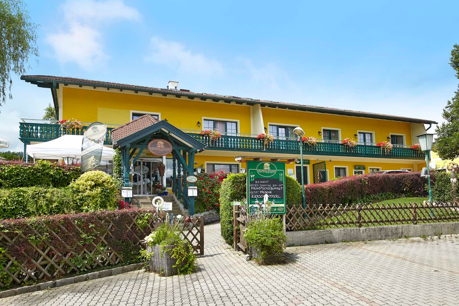 Gelbes Gebäude mit Balkon und Blumen, Krumbacherhof, Eingang mit Vordach und Schildern.