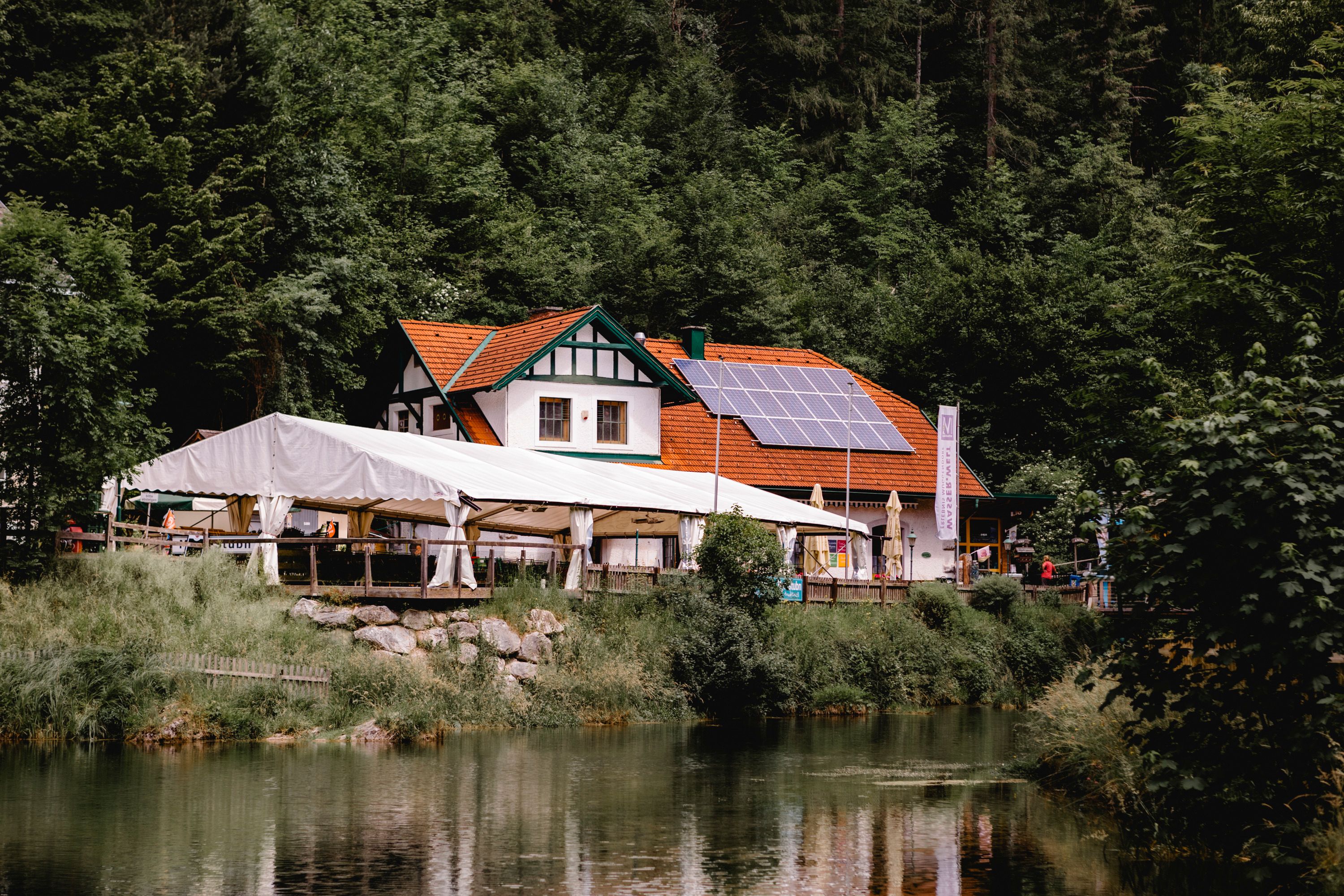 Gasthaus mit Solarpanelen und Zelt am Waldrand, nahe einem Teich.