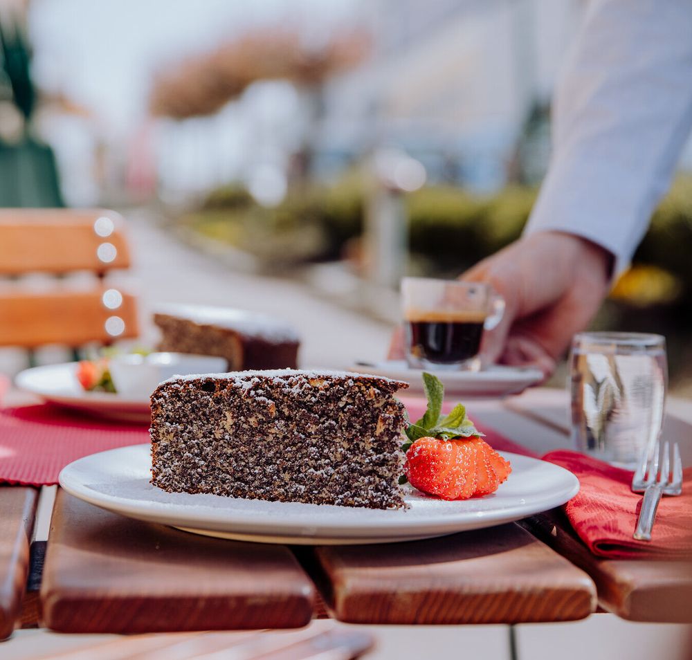 Genießen Sie die köstliche Waldviertler Mohntorte, die auf einem einladenden Tisch im Gastgarten serviert wird. Der Duft von frisch gebrühtem Kaffee mischt sich mit der warmen Sommerluft und schafft eine perfekte Atmosphäre für entspannte Stunden im Freien.
