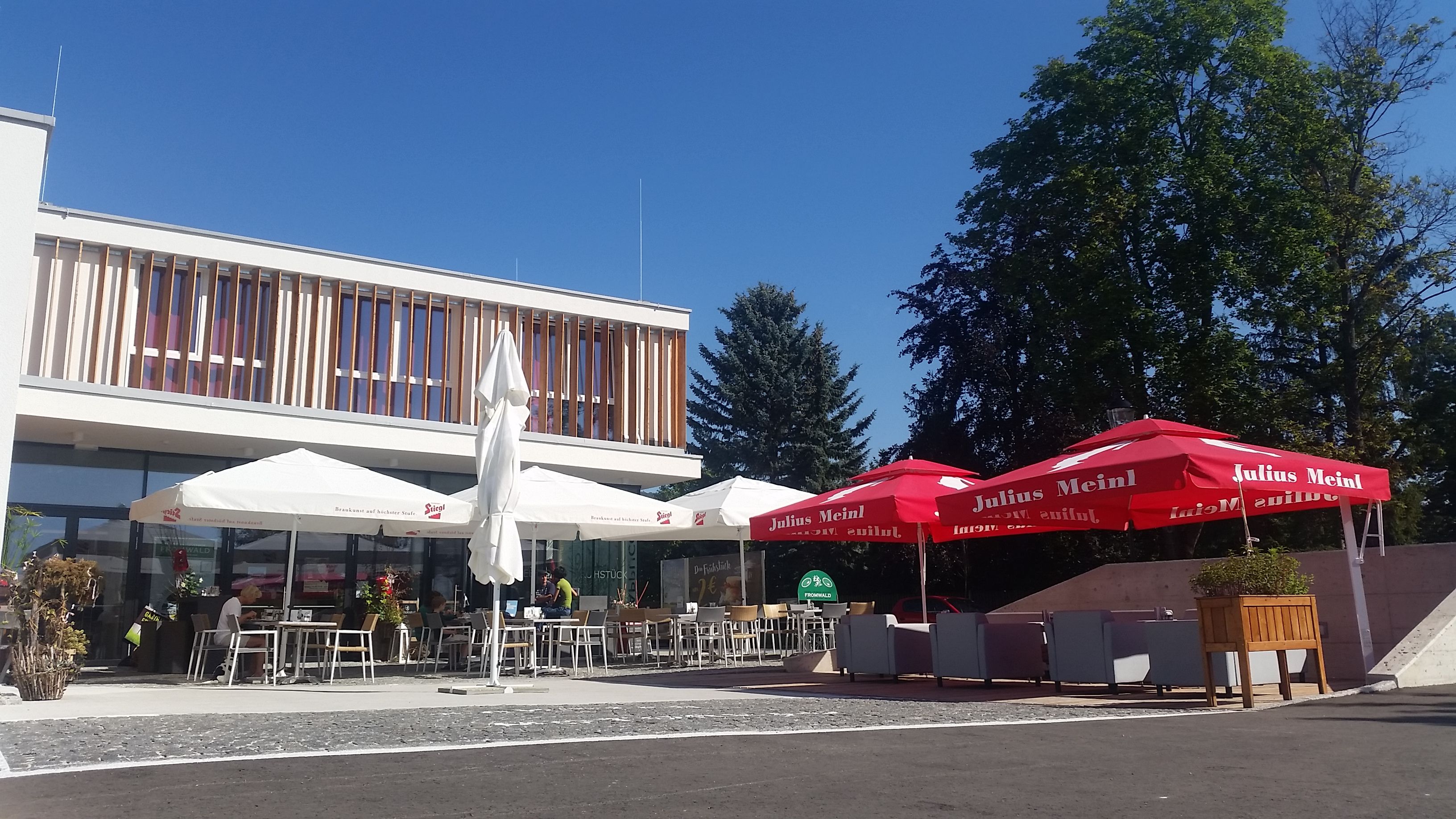 Ein moderner Gastgarten mit Sonnenschirmen und Sitzgelegenheiten vor einem Gebäude bei sonnigem Wetter.