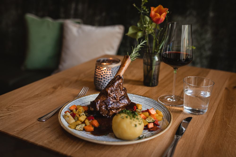 Ein Teller mit Lammstelze, Knödel und Gemüse, daneben ein Glas Rotwein und Wasser auf einem Holztisch.