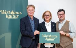 Drei Personen stehen vor einem Roll-up der Wirtshauskultur Niederösterreich und halten gemeinsam ein Schild mit dem Logo der Wirtshauskultur in die Kamera.