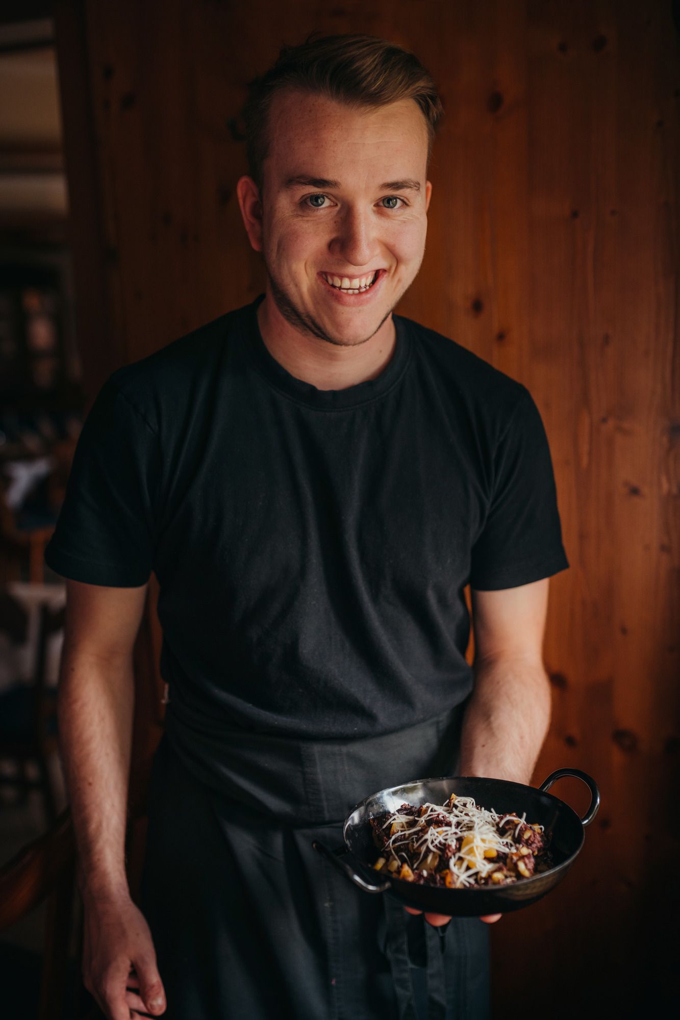 Ein lächelnder Mann in schwarzem T-Shirt hält eine Pfanne mit Essen.