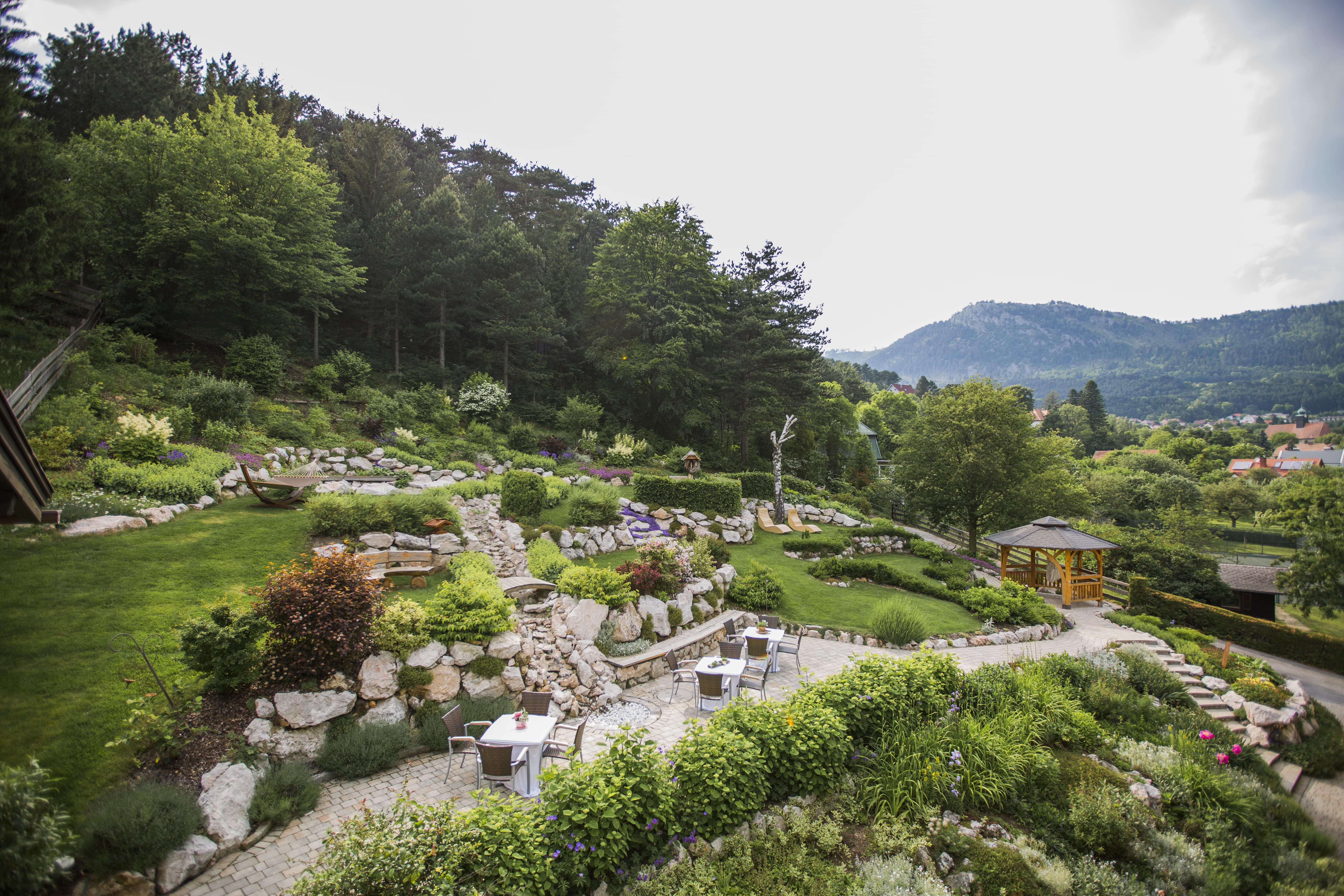 Ein terrassierter Garten mit Steinen, Pflanzen und Sitzbereichen, umgeben von Bäumen und Bergen im Hintergrund.