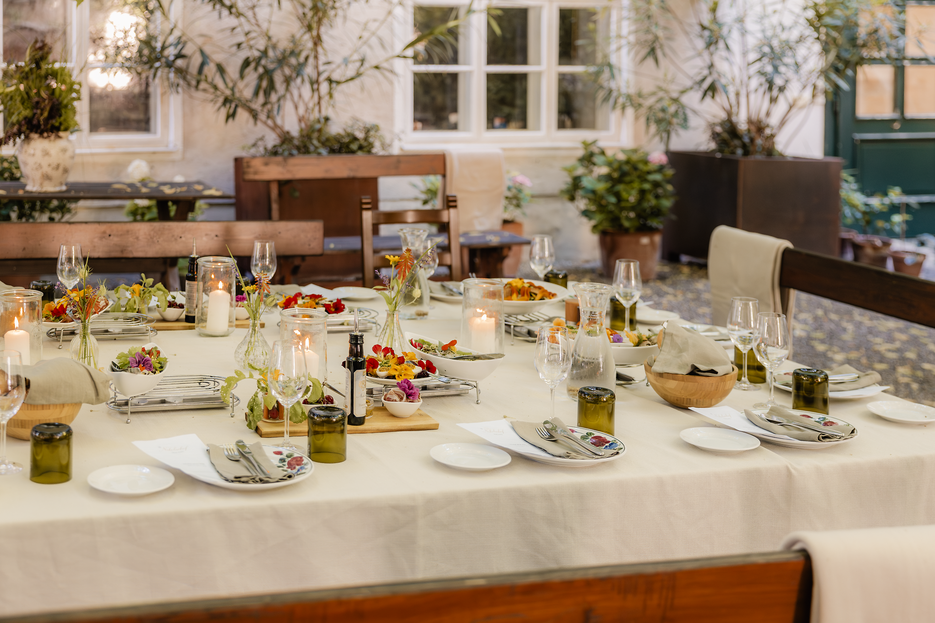 Großer gedeckter Tisch mit Tellern, Gläsern, Kerzen, Blumenvasen und mehreren angerichteten Speisen in einem begrünten Innenhof. Im Hintergrund sind Sitzplätze und Fenster zu sehen.