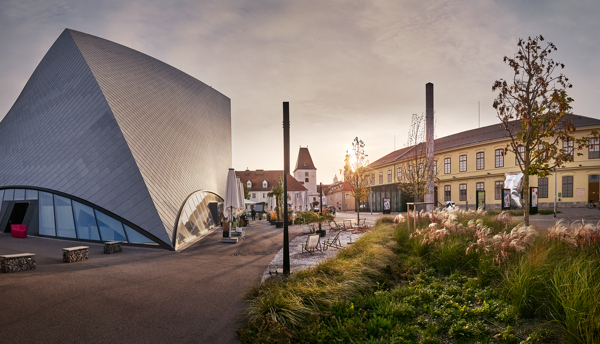 Die harmonische Verbindung von moderner Architektur und historischer Kulisse schafft eine inspirierende Atmosphäre. Sanfte Lichtstrahlen brechen durch die Wolken und tauchen den Platz in ein warmes Licht, während die umliegenden Bäume und Pflanzen eine einladende Ruhe ausstrahlen.