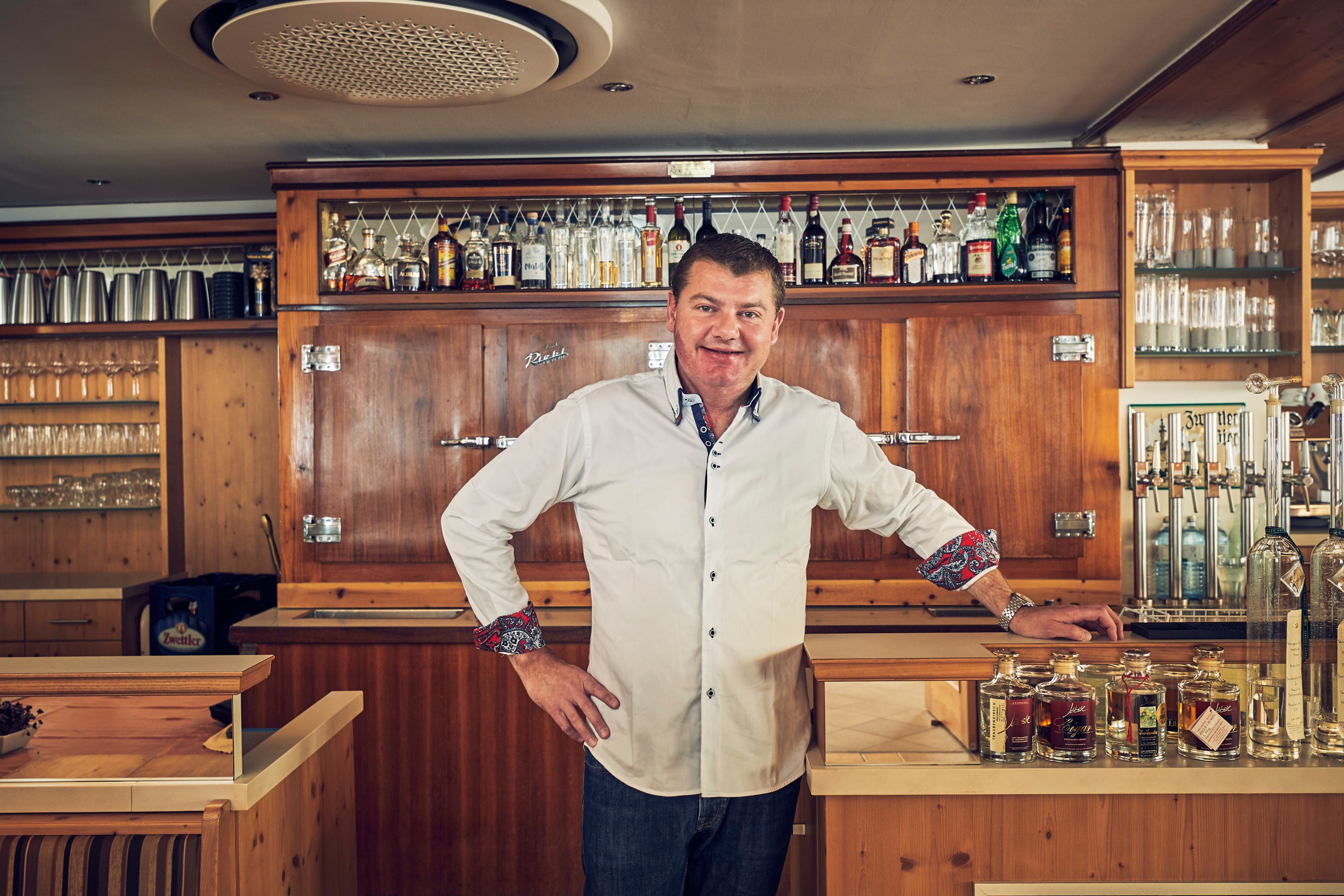 Ein Mann in einem weißen Hemd steht hinter einer Bar mit Flaschen und Gläsern im Hintergrund.