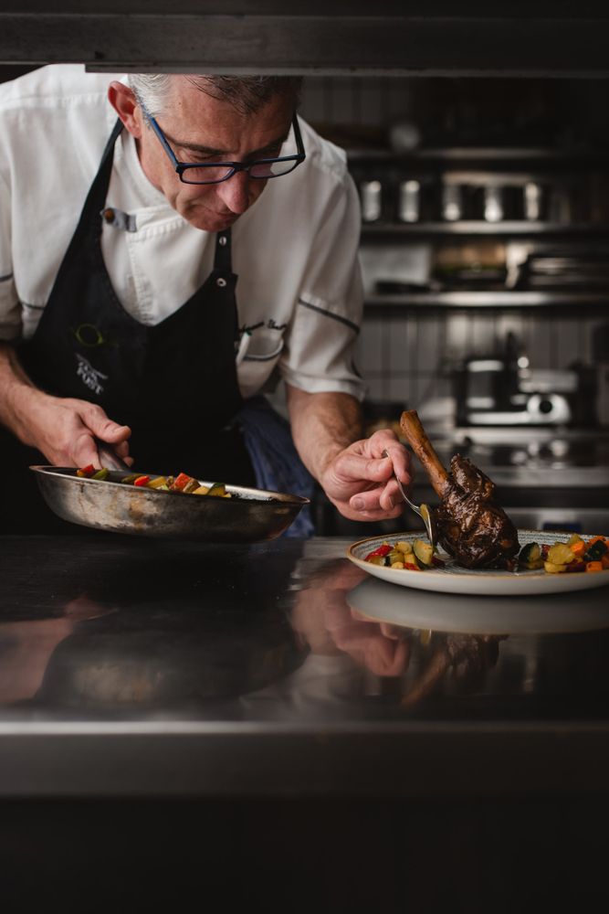 Ein Koch in einer professionellen Küche richtet ein Gericht mit Fleisch und Gemüse auf einem Teller an.