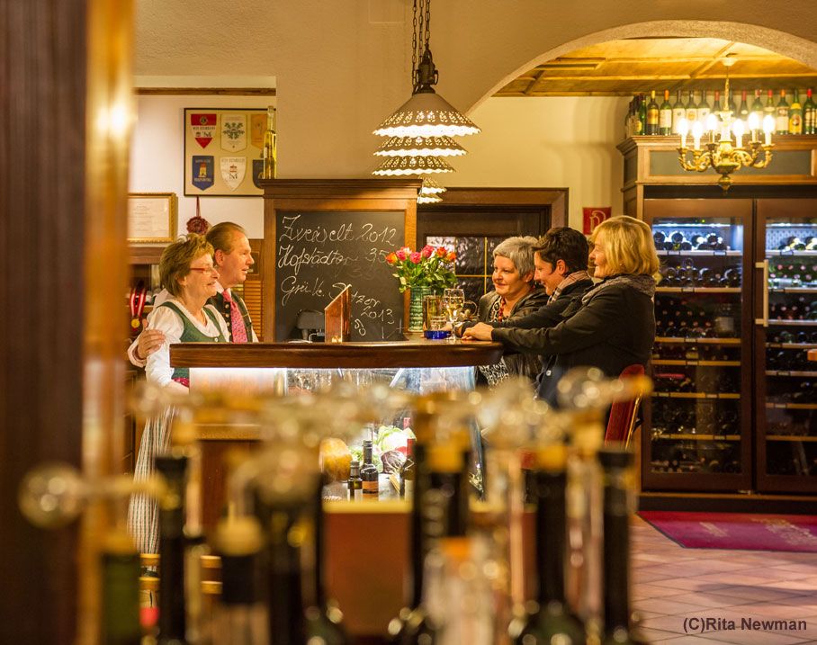 Menschen in einer gemütlichen Gaststube mit Weinflaschen im Hintergrund.