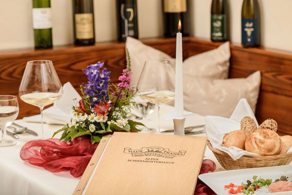 Eleganter gedeckter Tisch mit Speisekarte, Kerze, Blumen und Weinflaschen im Hintergrund.