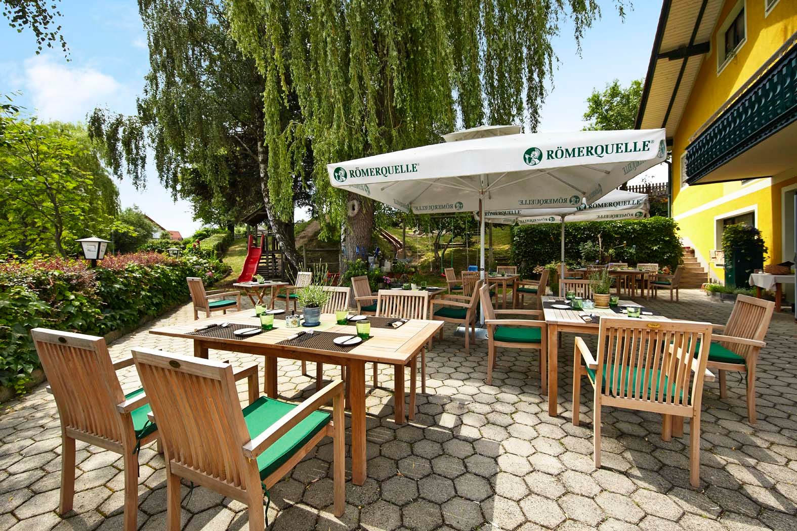Ein sonniger Gastgarten mit Holzmöbeln und Sonnenschirmen, umgeben von grüner Vegetation und einem gelben Gebäude.