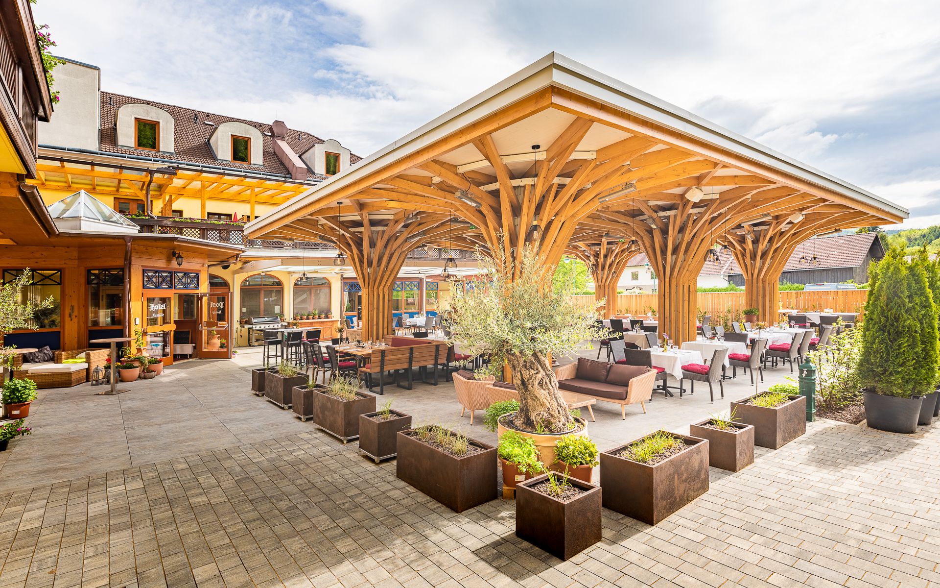 Terrasse mit Holzdach und Tischen, umgeben von Pflanzen.