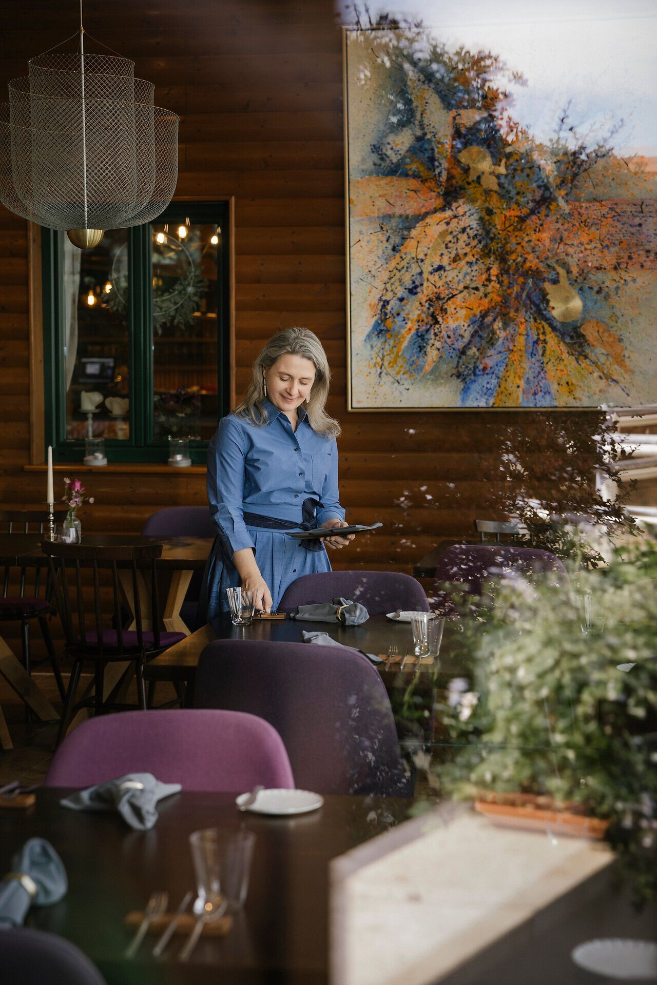 Eine Person deckt einen Tisch im Innenbereich eines Restaurants. Auf den Tischen stehen Gläser, Geschirr und Blumenvasen, im Hintergrund hängen große Bilder an der Wand.