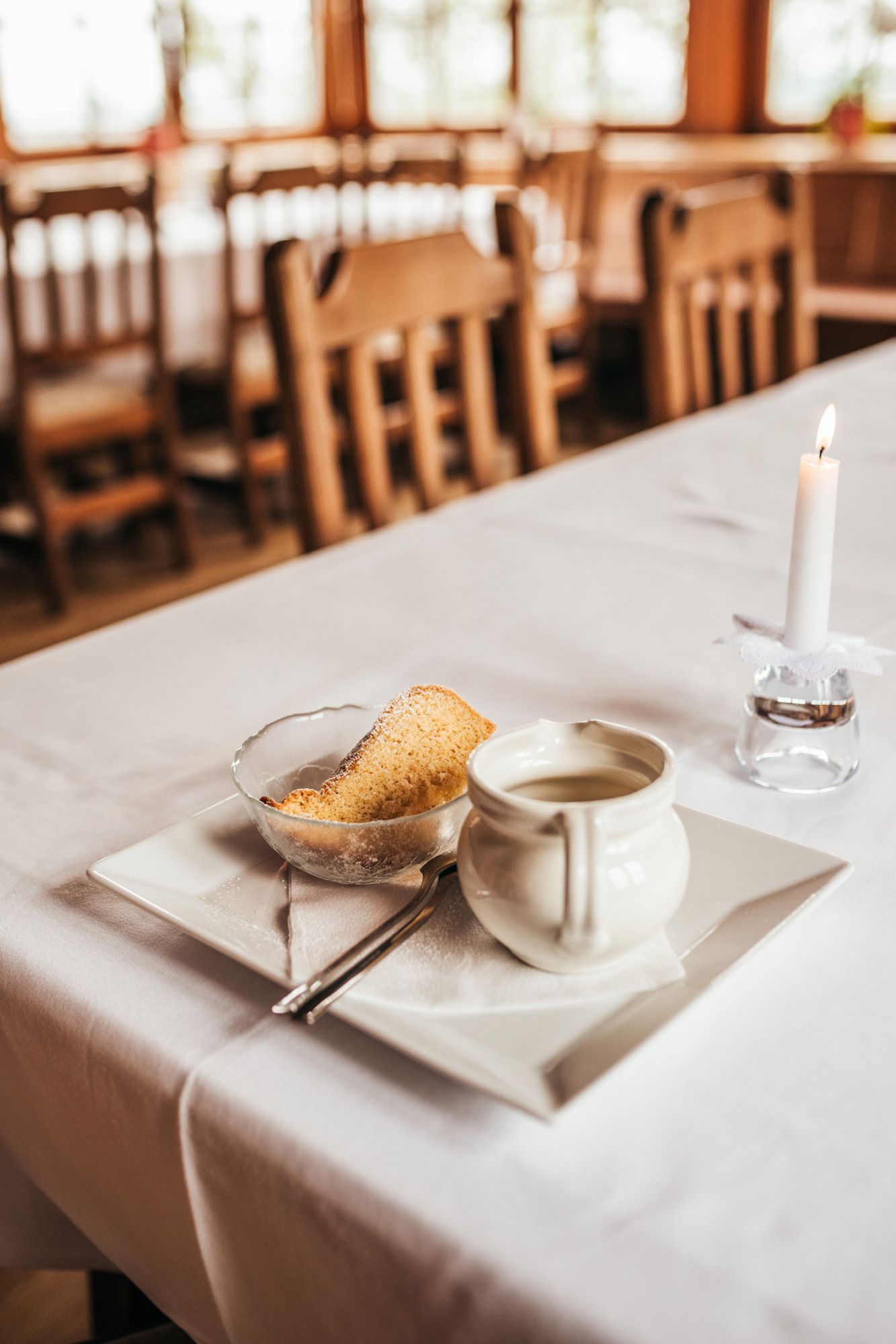 A laid table with a plate, a cup, a piece of cake and a lit candle.