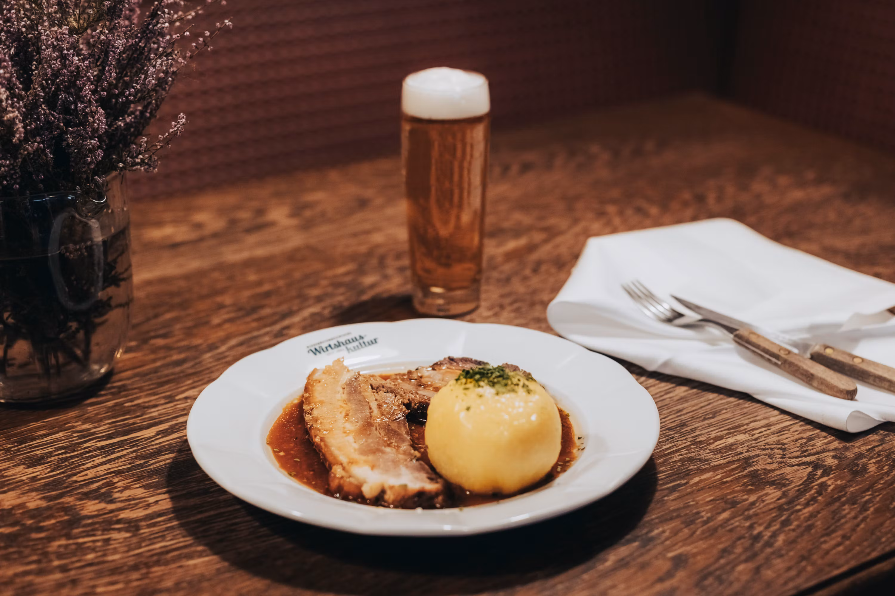 Ein Teller mit Schweinsbraten und Waldviertler Knödel, daneben ein Glas Bier auf einem Holztisch.
