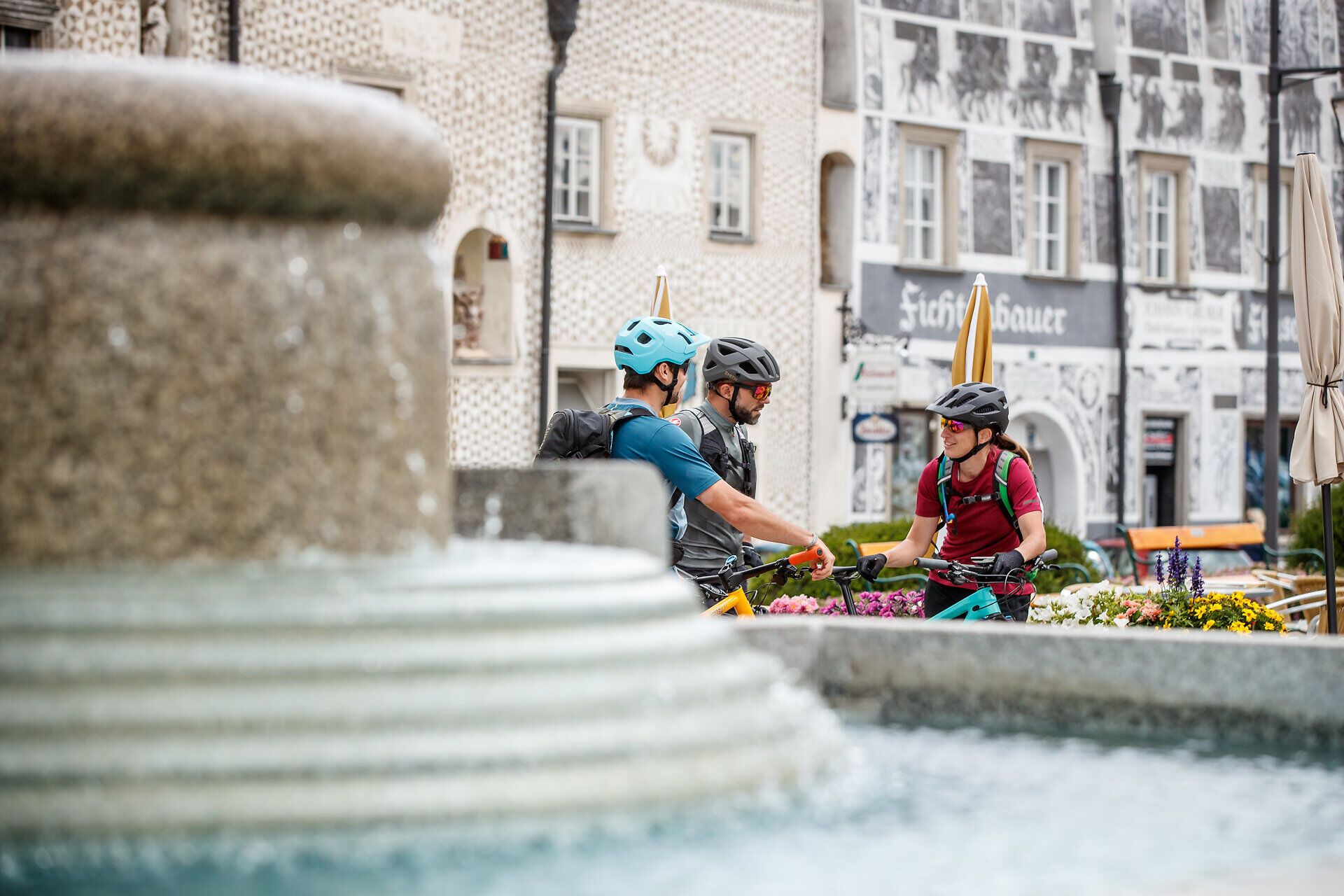 Drei Radfahrer genießen eine entspannte Pause am Stadtplatz, umgeben von blühenden Blumen und der charmanten Architektur. Die Atmosphäre ist lebhaft und einladend, während die frische Luft und die Natur zum Erkunden einladen.