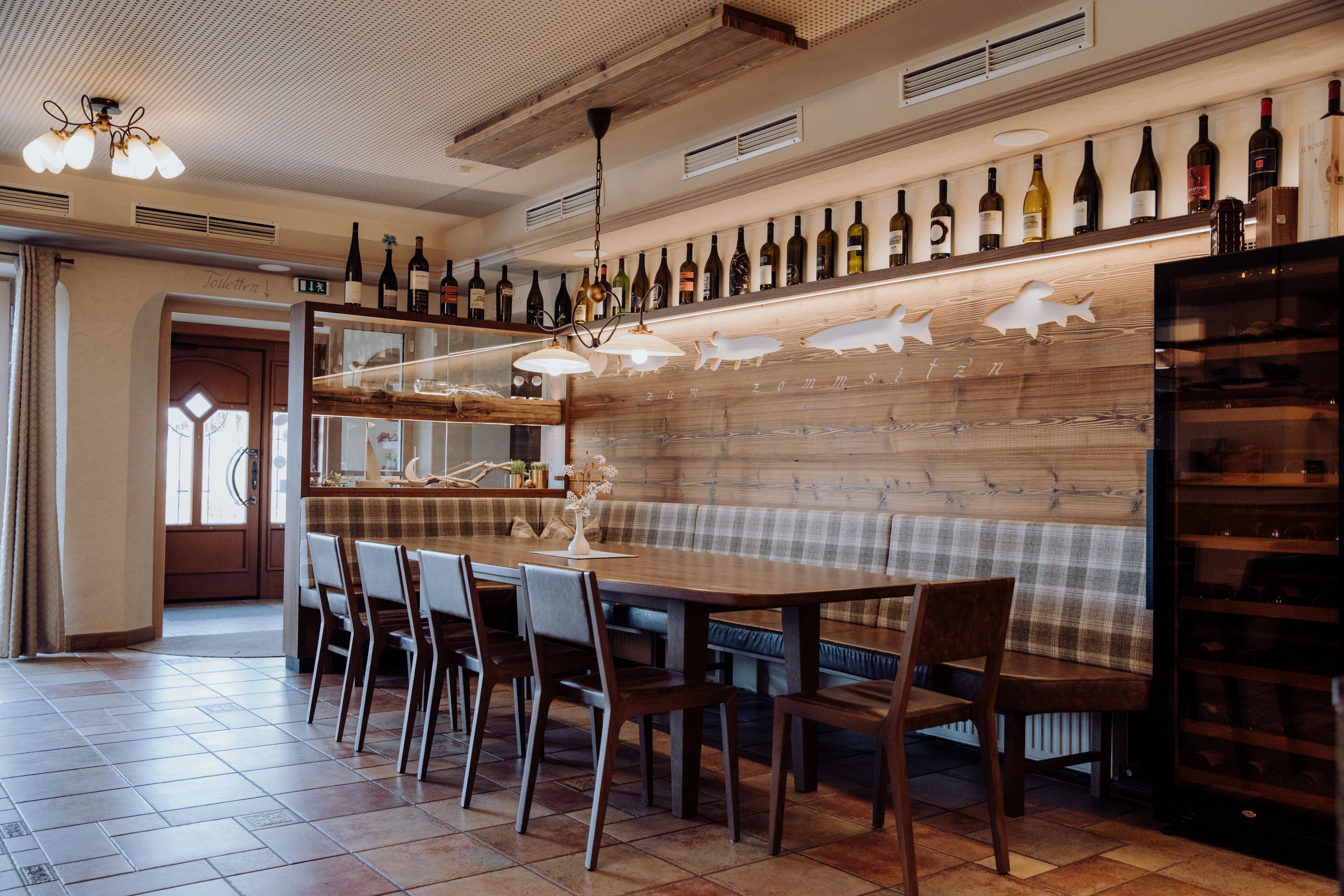 Gemütlicher Essbereich mit Holztisch, Stühlen und Bank, dekoriert mit Weinflaschen und Fischmotiven an der Wand.