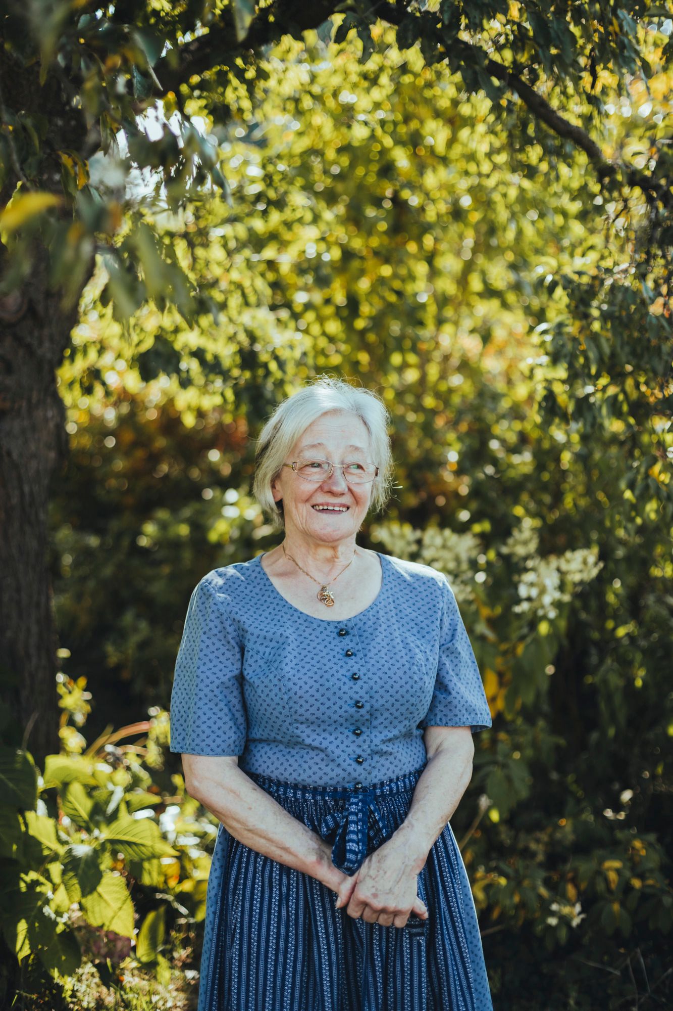 Ältere Frau in blauem Kleid steht lächelnd im Garten.