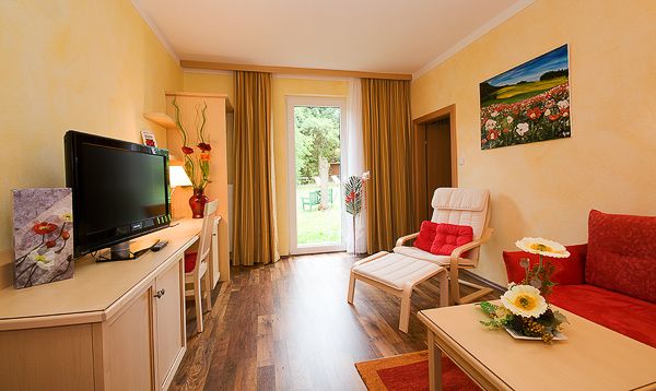 Gemütliches Zimmer mit roten Akzenten, Fernseher, Sofa, Sessel und Blick auf den Garten durch eine Glastür.