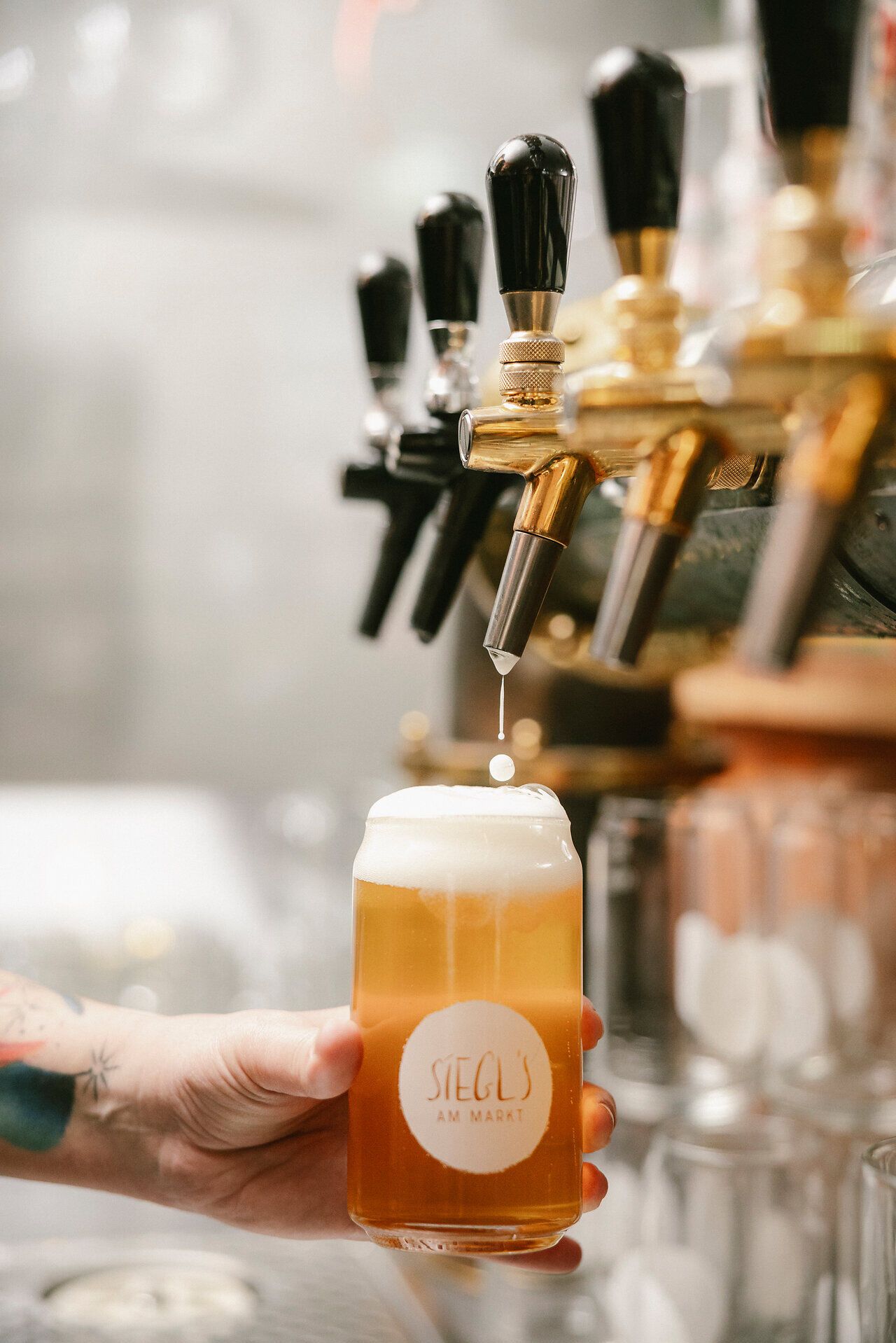 Vom Zapfhahn tropft frisch gezapftes Bier in ein volles Glas mit der Aufschrift "Siegl´s am Markt", das von einer Hand gehalten wird.