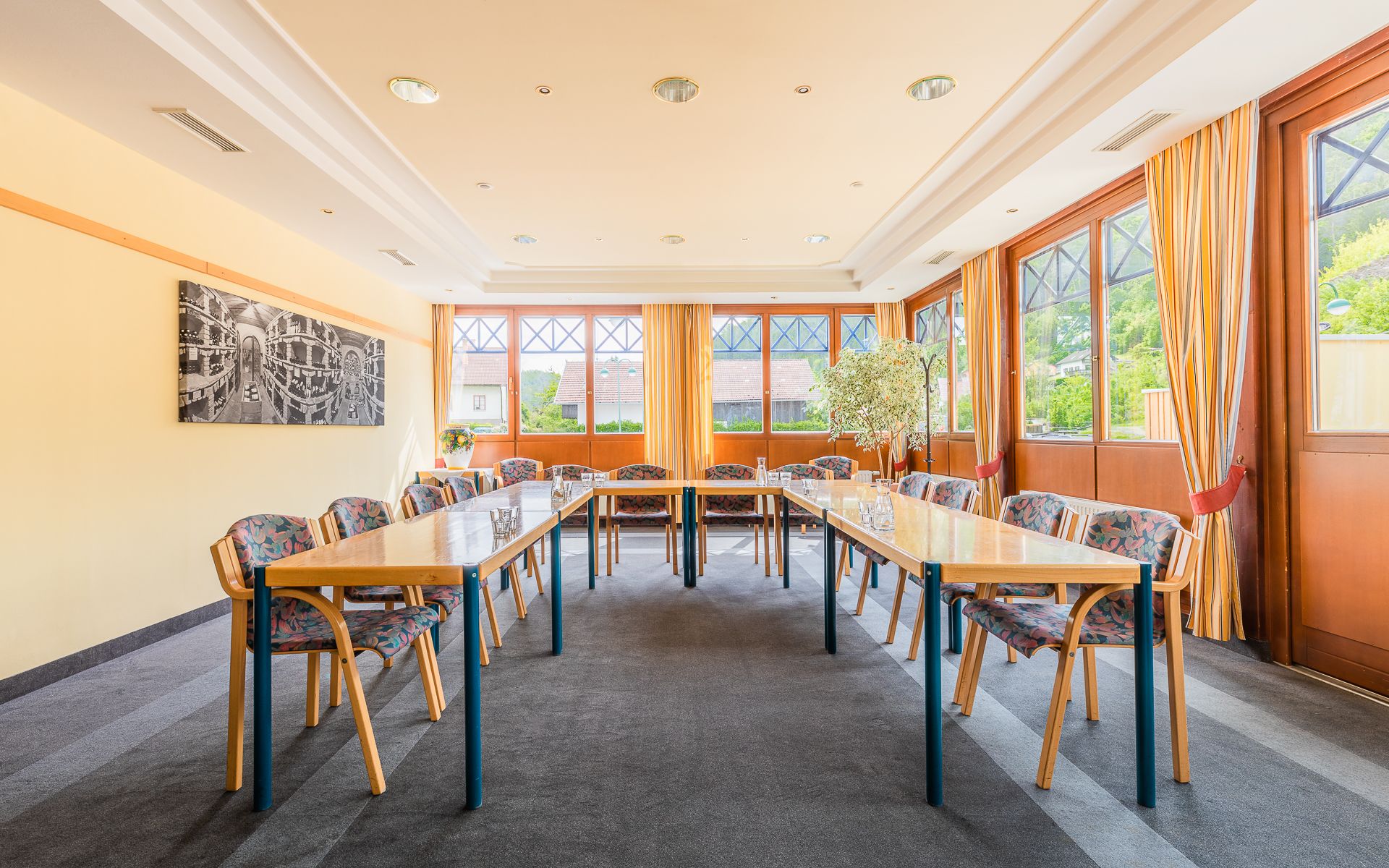 Ein heller Seminarraum mit Tischen in U-Form, Stühlen und großen Fenstern mit Vorhängen.