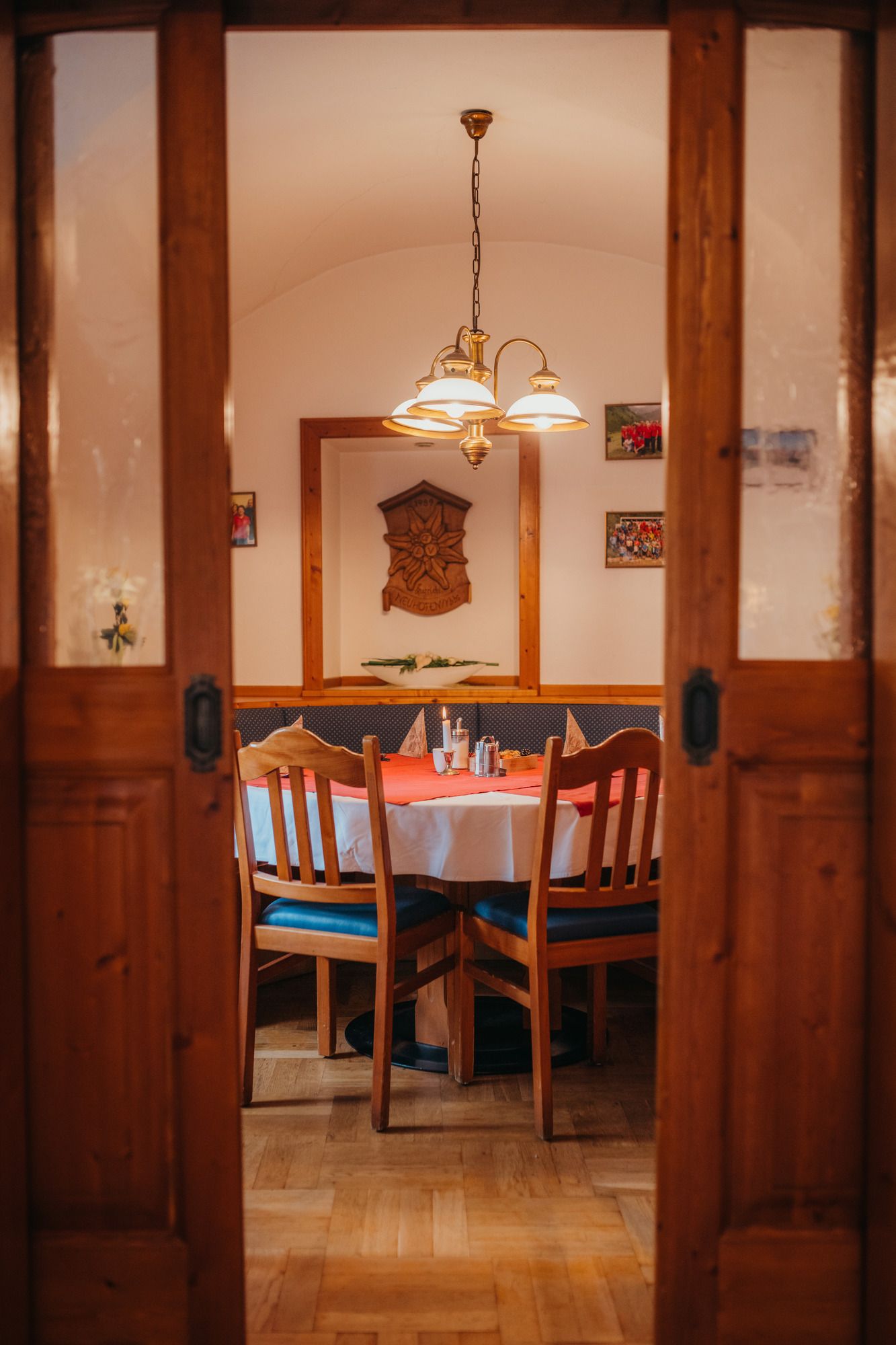 Ein gemütlicher Raum mit Holzmöbeln, einem runden Tisch mit rotem Tischtuch und einer Hängelampe. An der Wand hängen Bilder und ein Holzrelief.