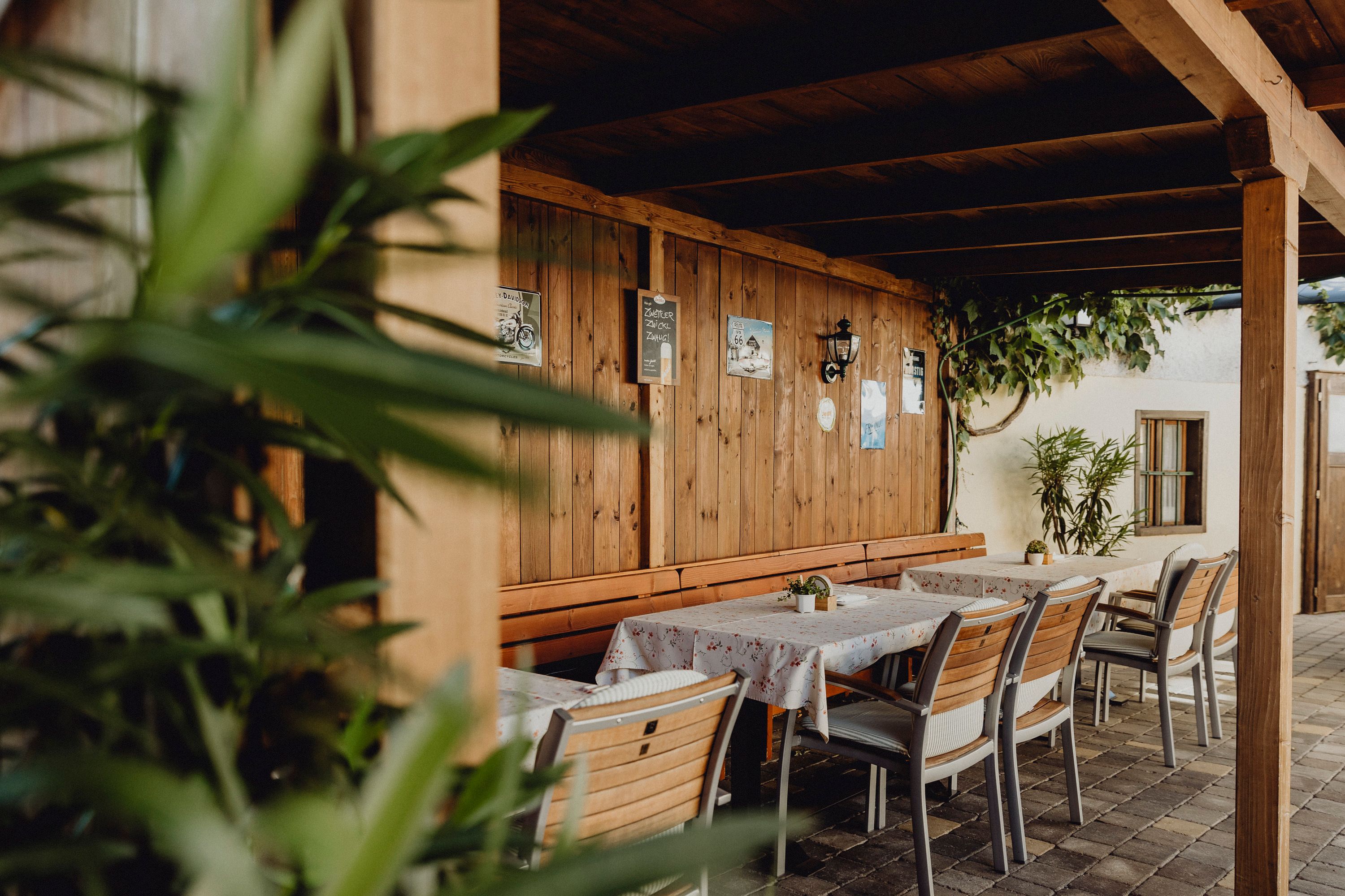 Überdachter Gastgarten mit Holzwänden, Tischen und Stühlen, dekoriert mit Pflanzen und Postern.