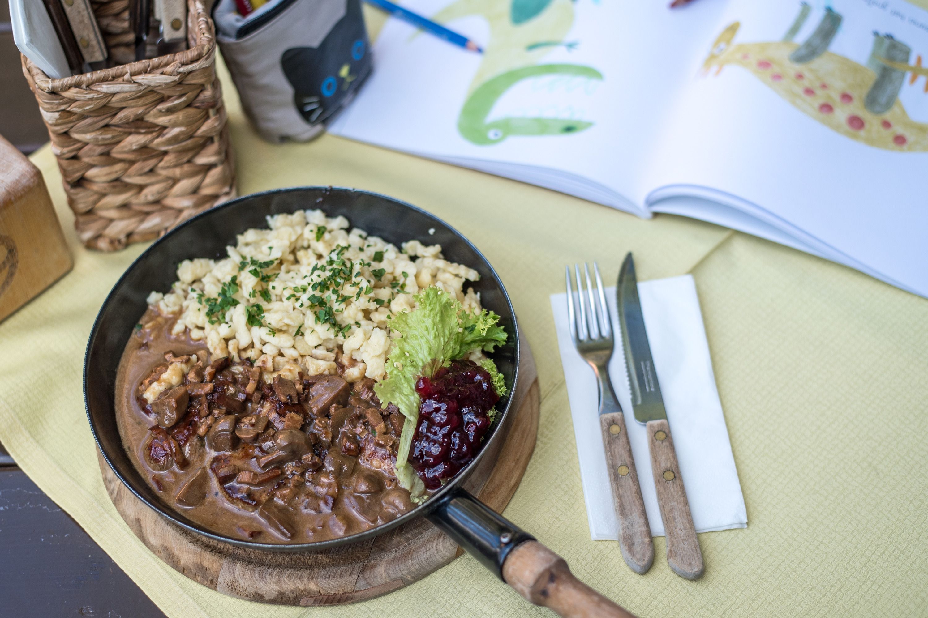 Ein Teller mit Spätzle, Fleisch und Preiselbeeren auf einem Tisch mit Besteck und einem offenen Buch.