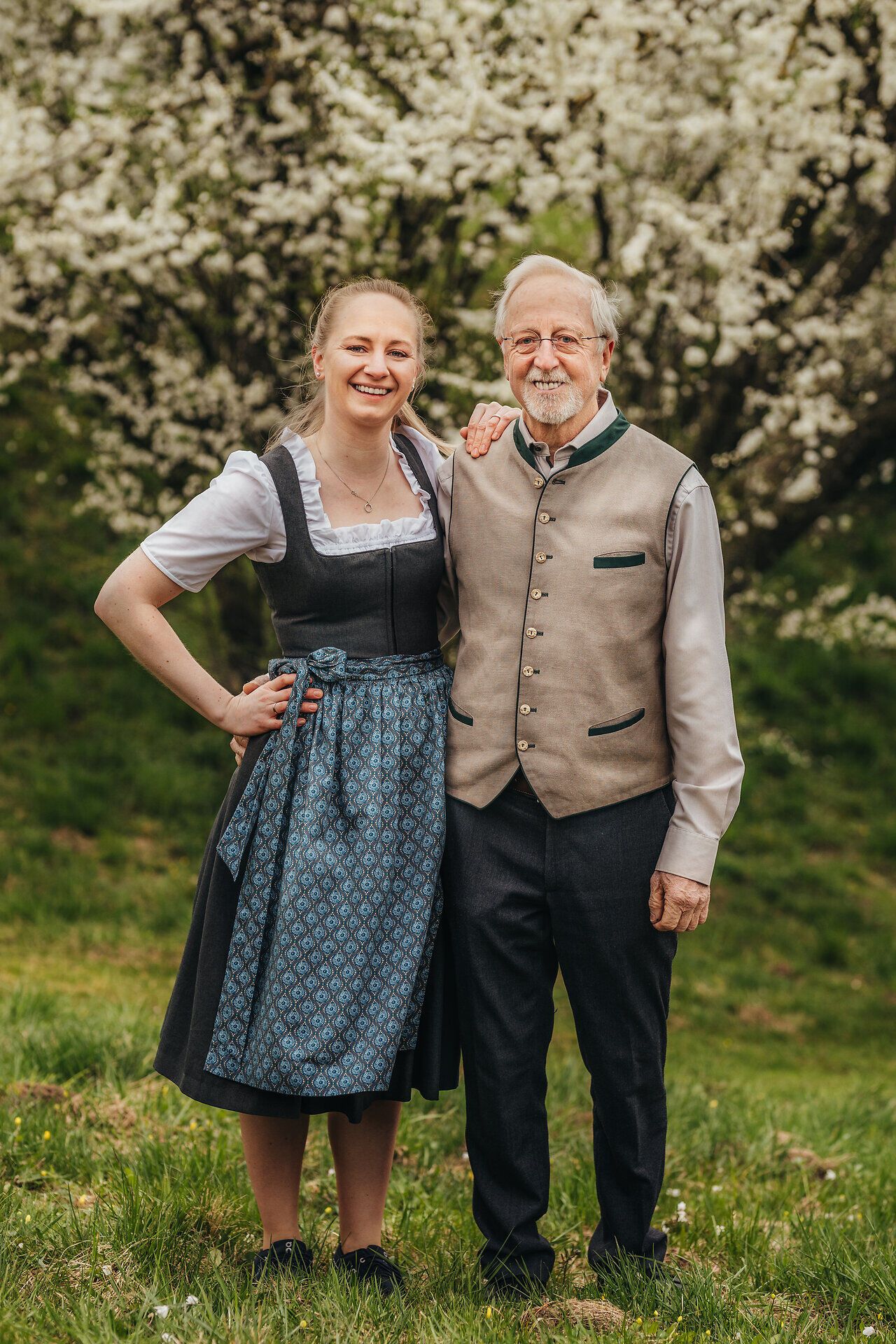 Inmitten blühender Obstbäume strahlen die fröhlichen Gesichter von Wirtin Maria Bachler und ihrem Begleiter. Die harmonische Atmosphäre und die frische Luft laden dazu ein, die köstlichen Kräuter und den regionalen Most zu genießen, während die Natur in voller Pracht erblüht.
