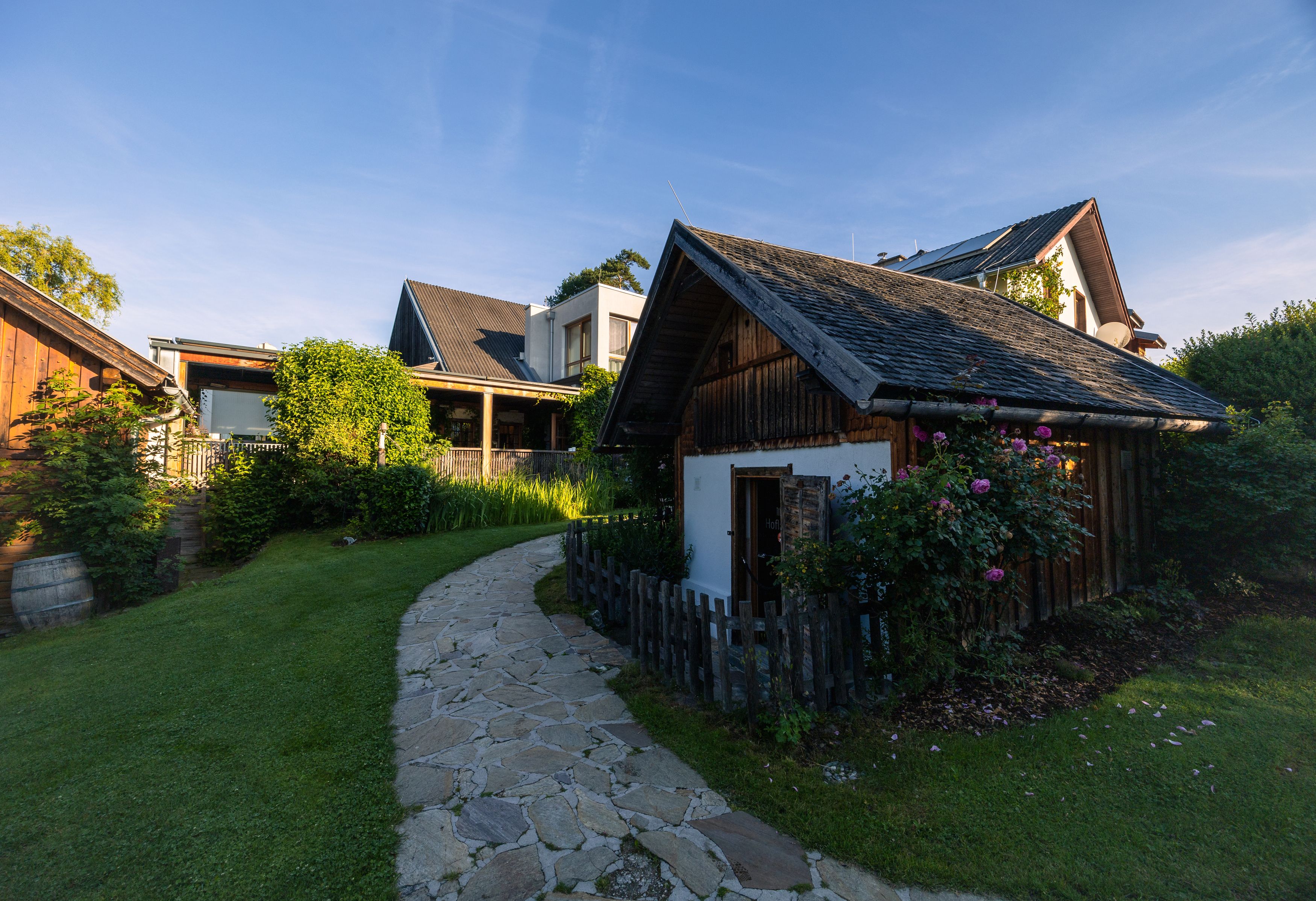 Ein kleiner Gartenweg führt zu einem traditionellen Holzhaus mit Blumen und Büschen, umgeben von weiteren Gebäuden unter blauem Himmel.