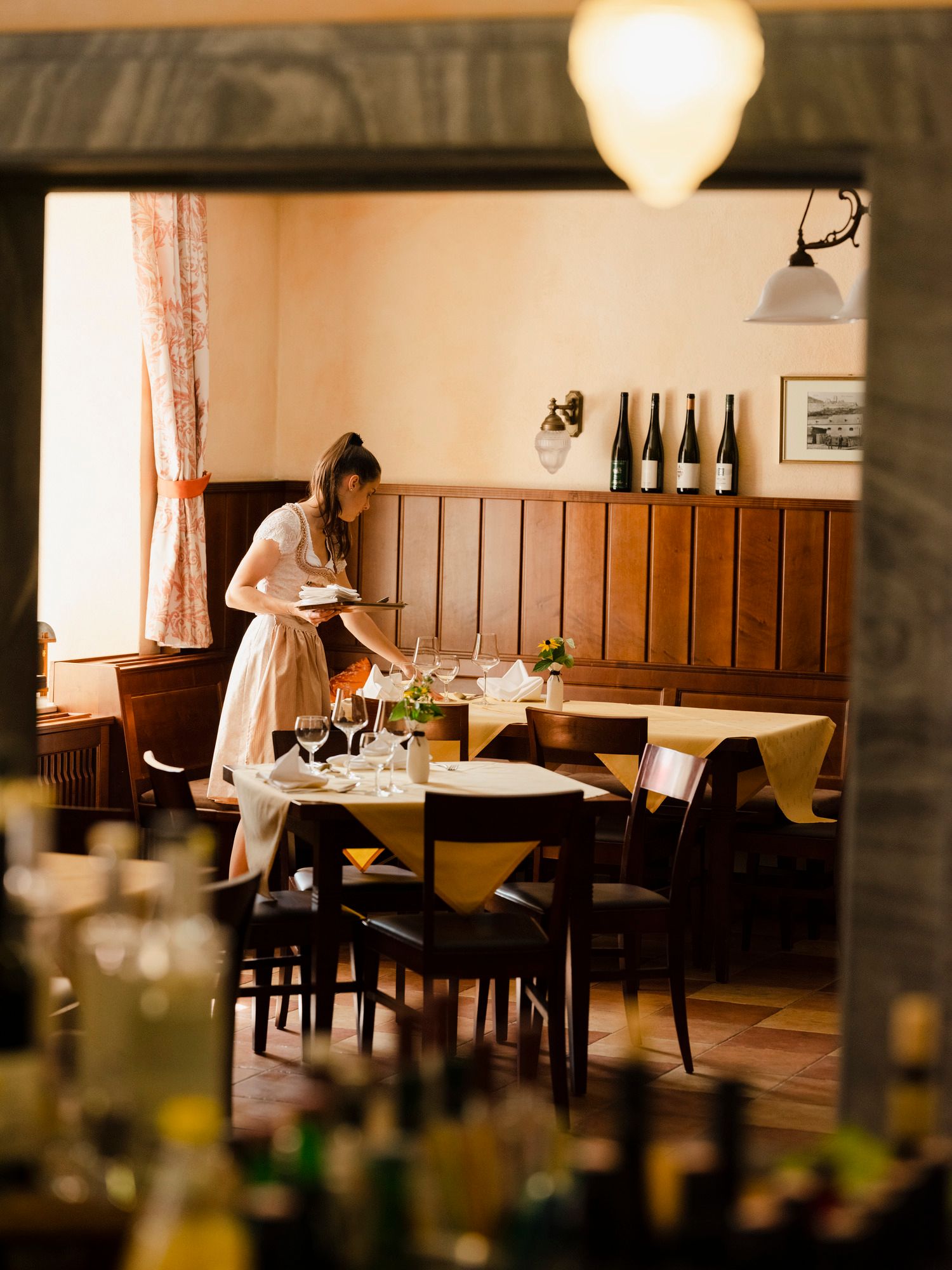 Eine Frau in traditioneller Kleidung deckt einen Tisch in einem gemütlichen Restaurant mit Holzwänden und Weinflaschen im Hintergrund.